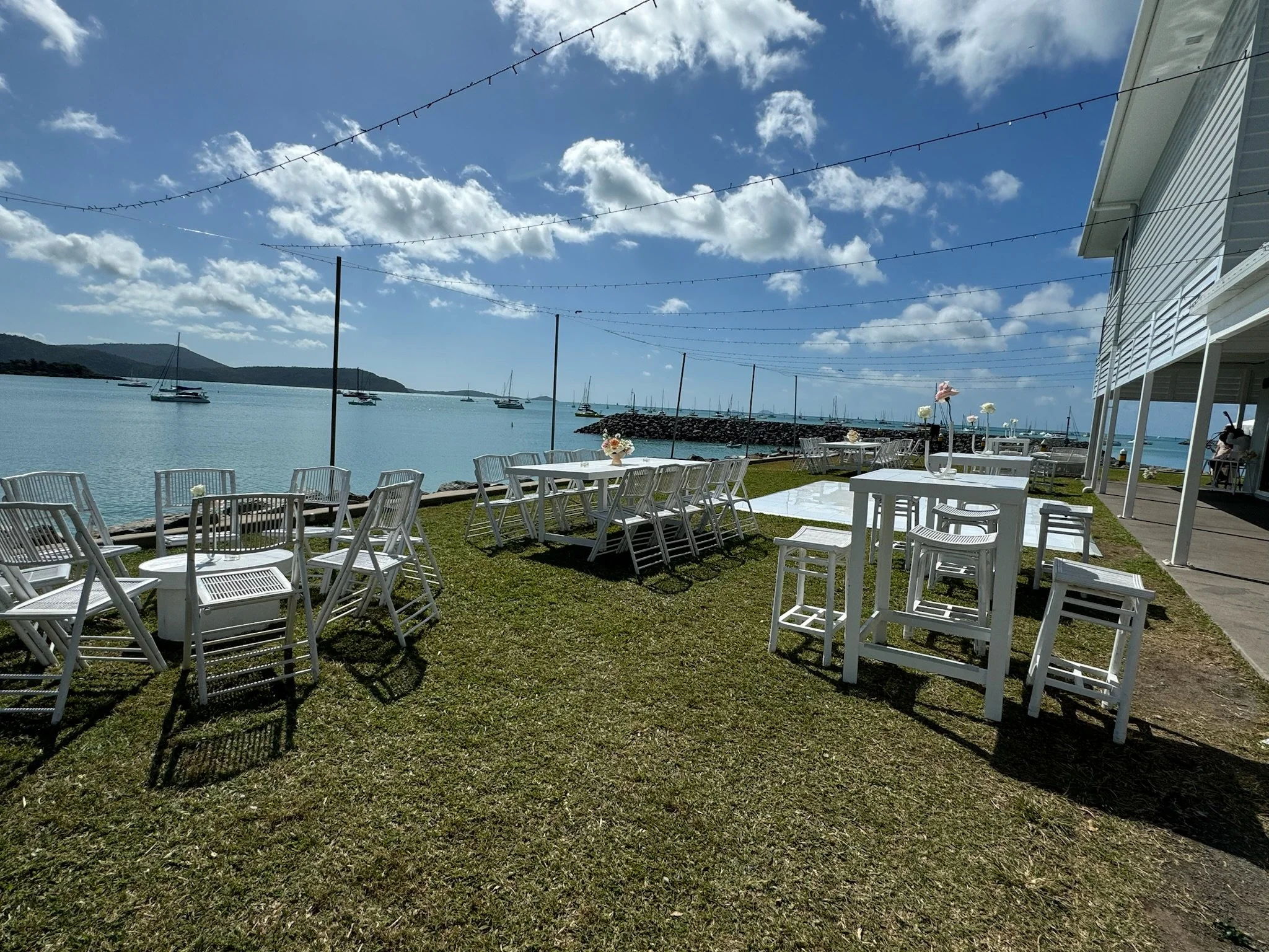 Outdoor seaside venue with white tables and chairs on grass, decorated with flowers, overlooking a calm harbor with anchored sailboats and a partly cloudy sky.