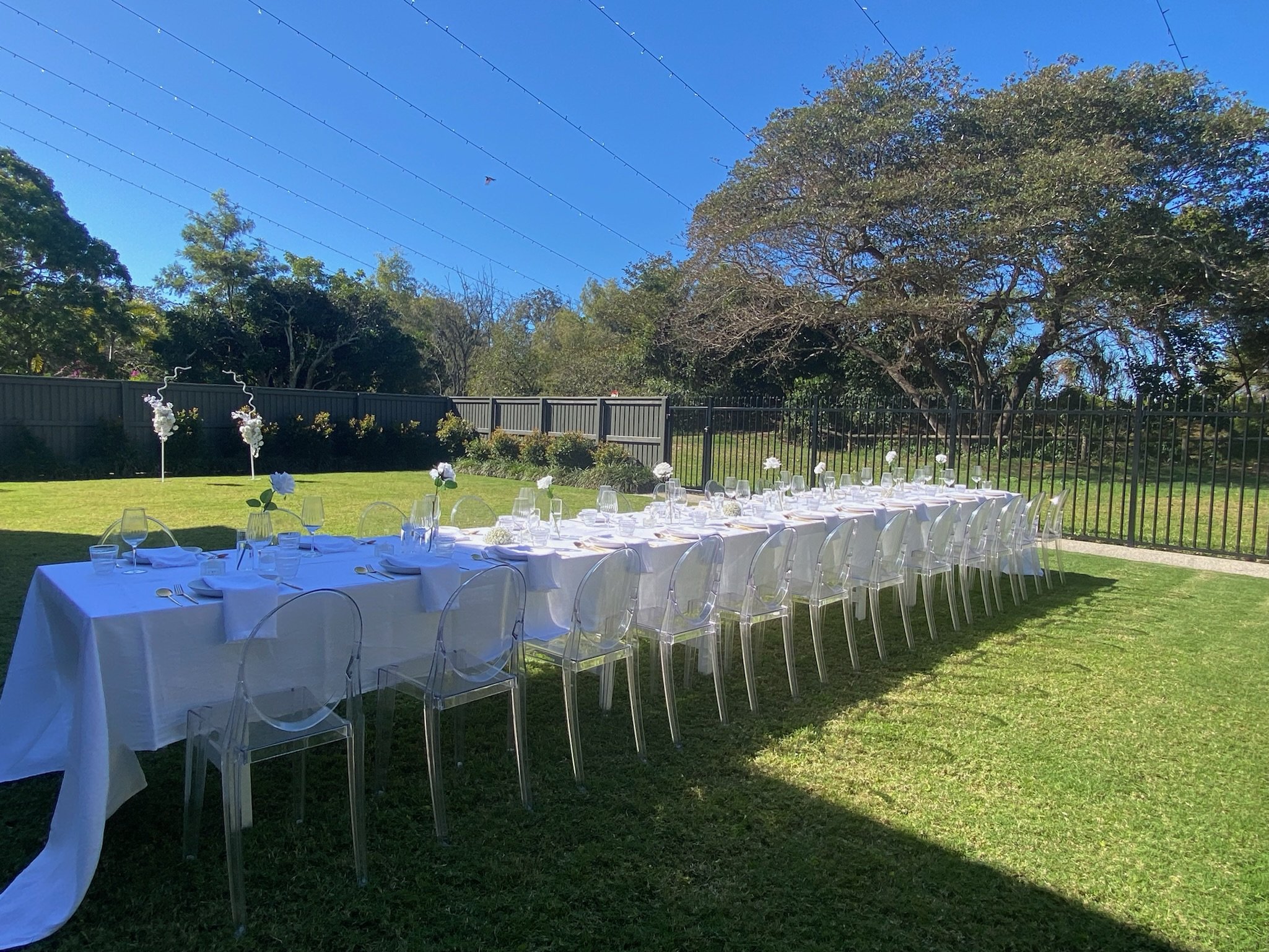 Long outdoor dining table with white tablecloth, set with wine glasses, plates, silverware, and small floral arrangements, on a grassy lawn under a clear blue sky, with trees and a fence in the background.