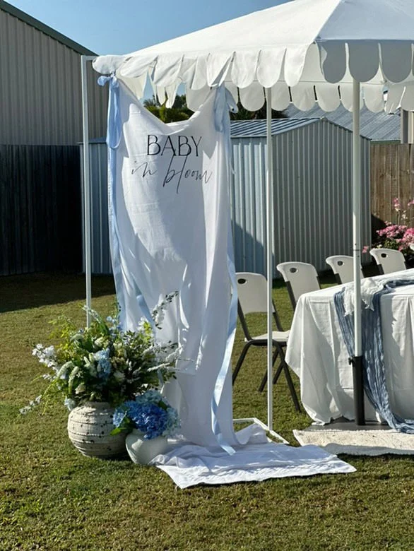 Outdoor baby shower setup with a white canopy, a large white fabric banner reading "Baby in Bloom," a floral arrangement with white and blue flowers in a textured white vase, and tables with white tablecloths and chairs on a grassy yard.