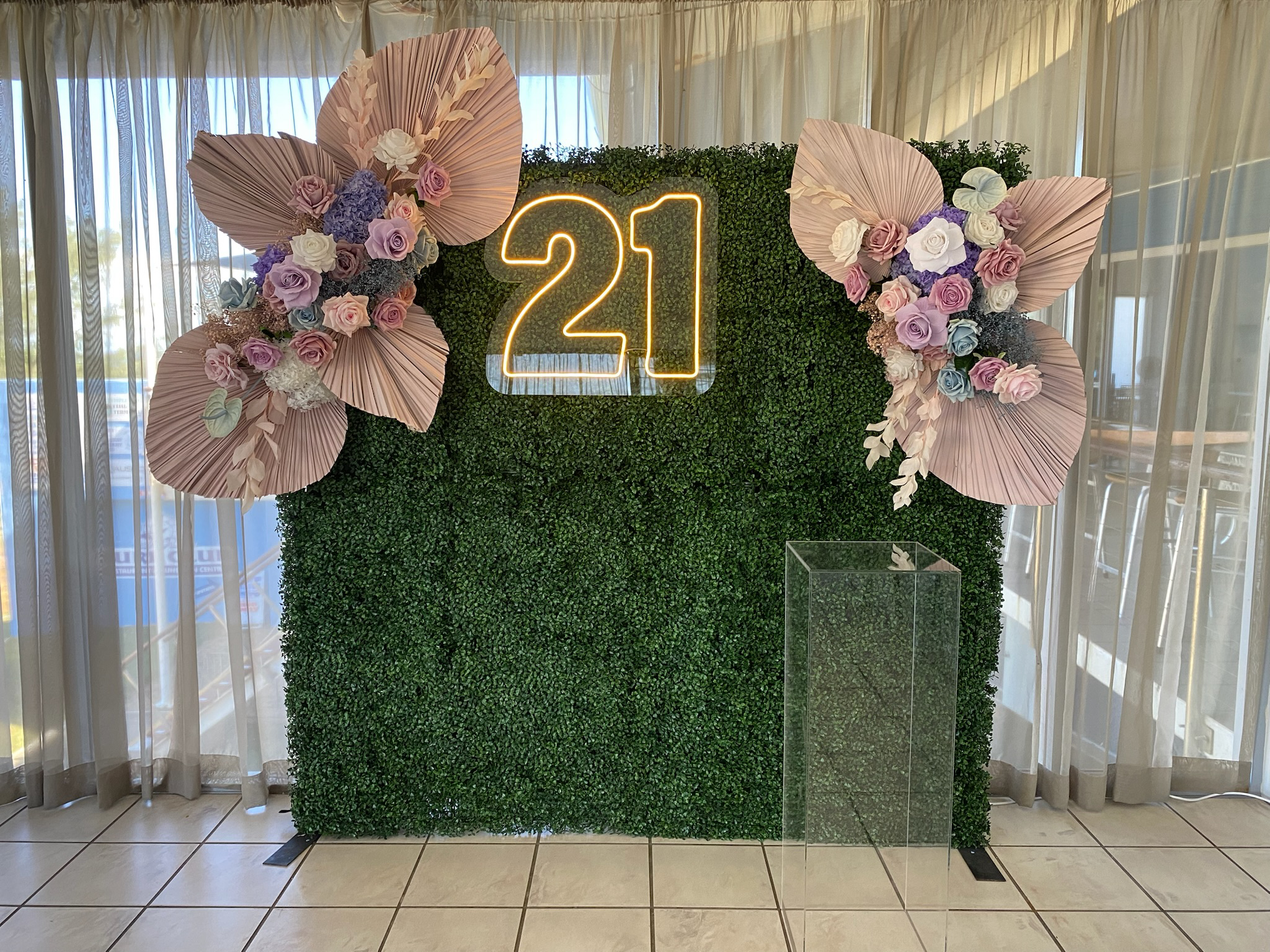 Decorative floral arrangement on a green hedge backdrop with a neon sign displaying the number '21', two large pink paper flowers, and colorful roses, placed indoors near windows with white curtains.