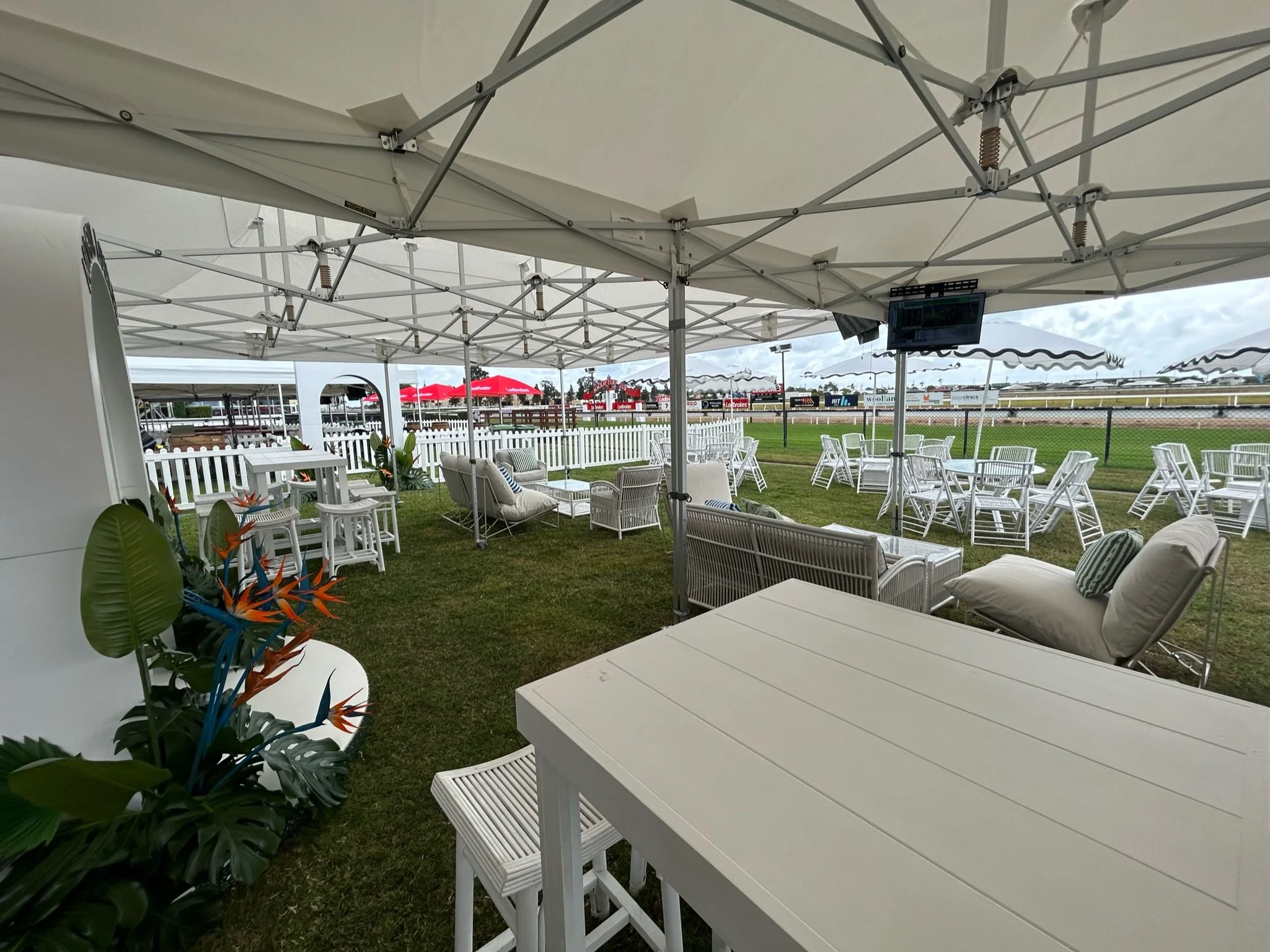 Outdoor patio area with white lounge chairs, tables, and umbrellas, overlooking a racetrack under a cloudy sky.