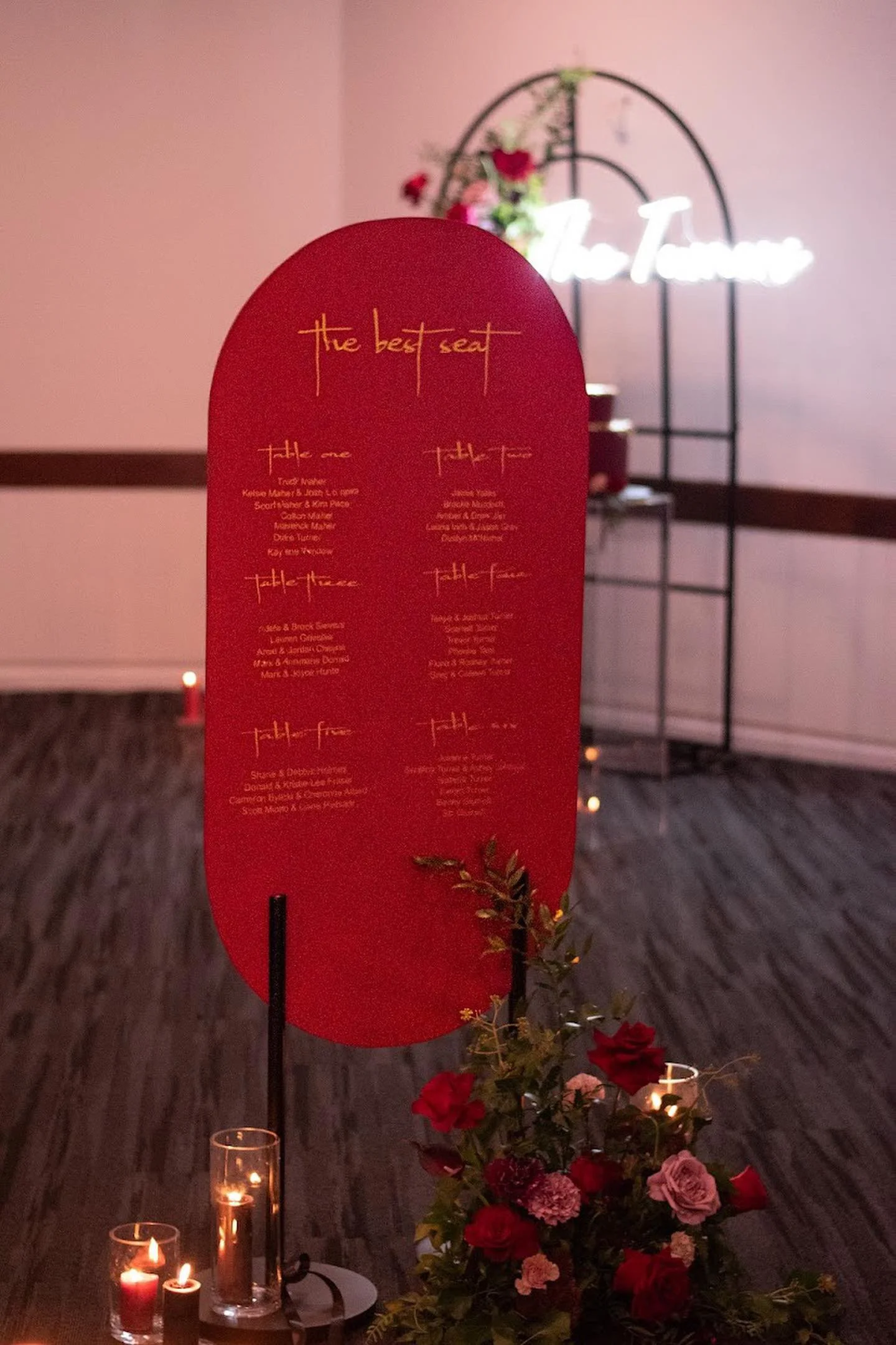 A red oval sign with the title 'the best seat' and a list of tables and names below, set in a dimly lit room with candles and flower arrangements, and a neon sign in the background.