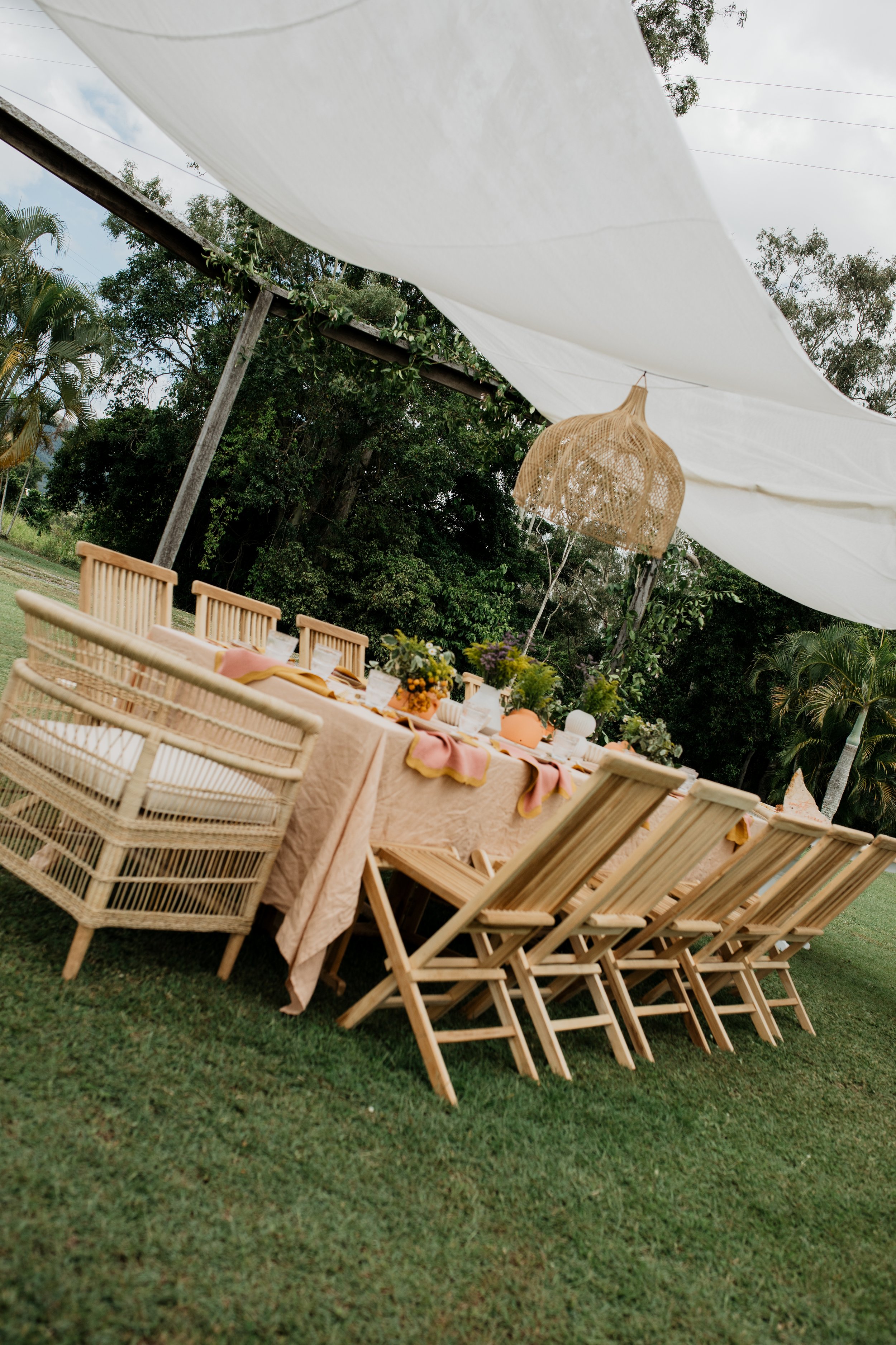 Outdoor dining setup with a long table covered with a pink dessert cloth, surrounded by wooden and wicker chairs, under a white canopy and hanging rattan pendant light, with greenery in the background.