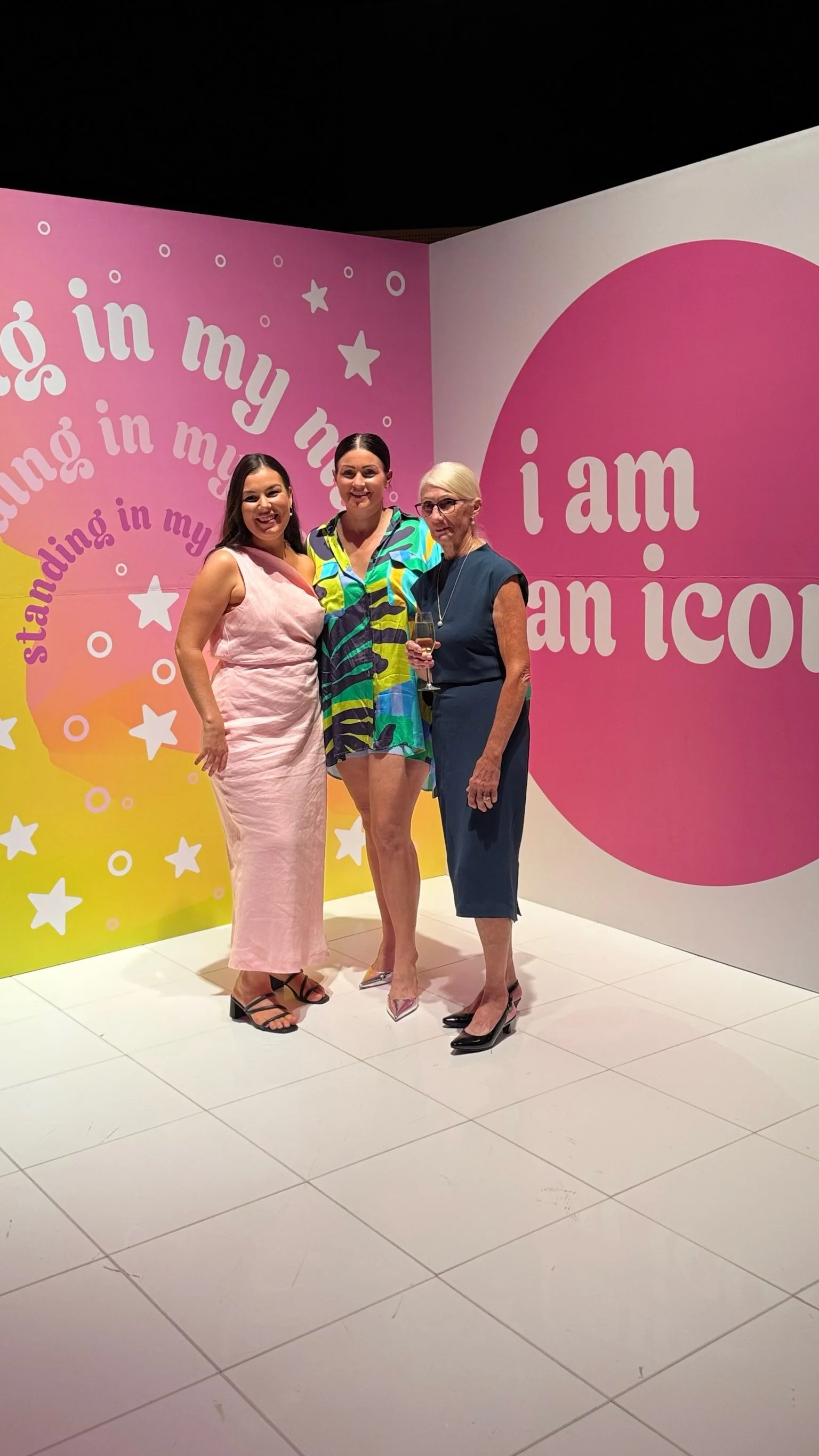 Three women standing together in front of a pink and yellow background with the text 'I am an icon' and 'stamping in my' repeated. They are dressed in stylish outfits and smiling.