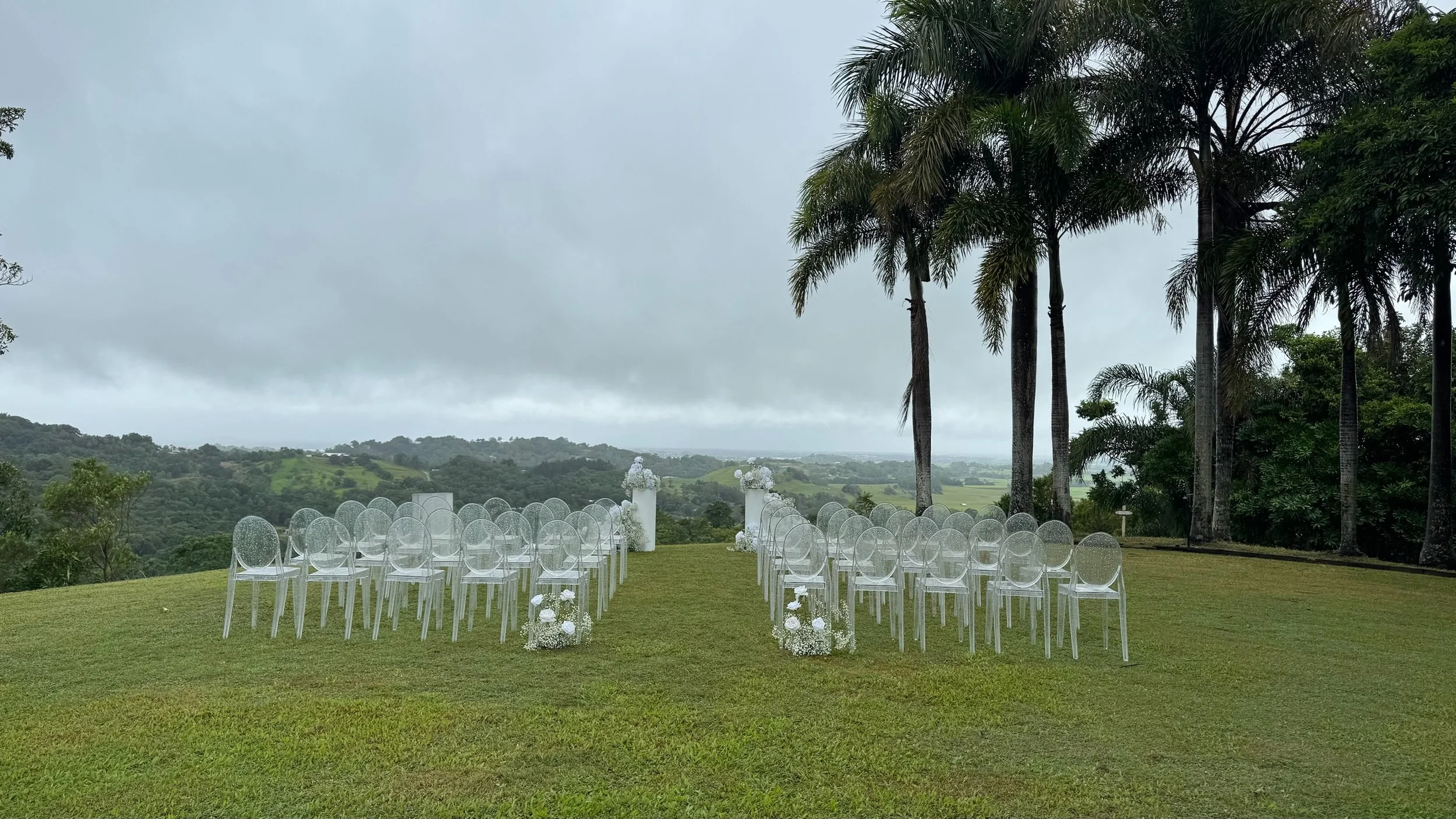 Outdoor wedding setup with transparent chairs arranged in rows on a green lawn, decorated with white floral arrangements, overlooking scenic hills under cloudy sky.