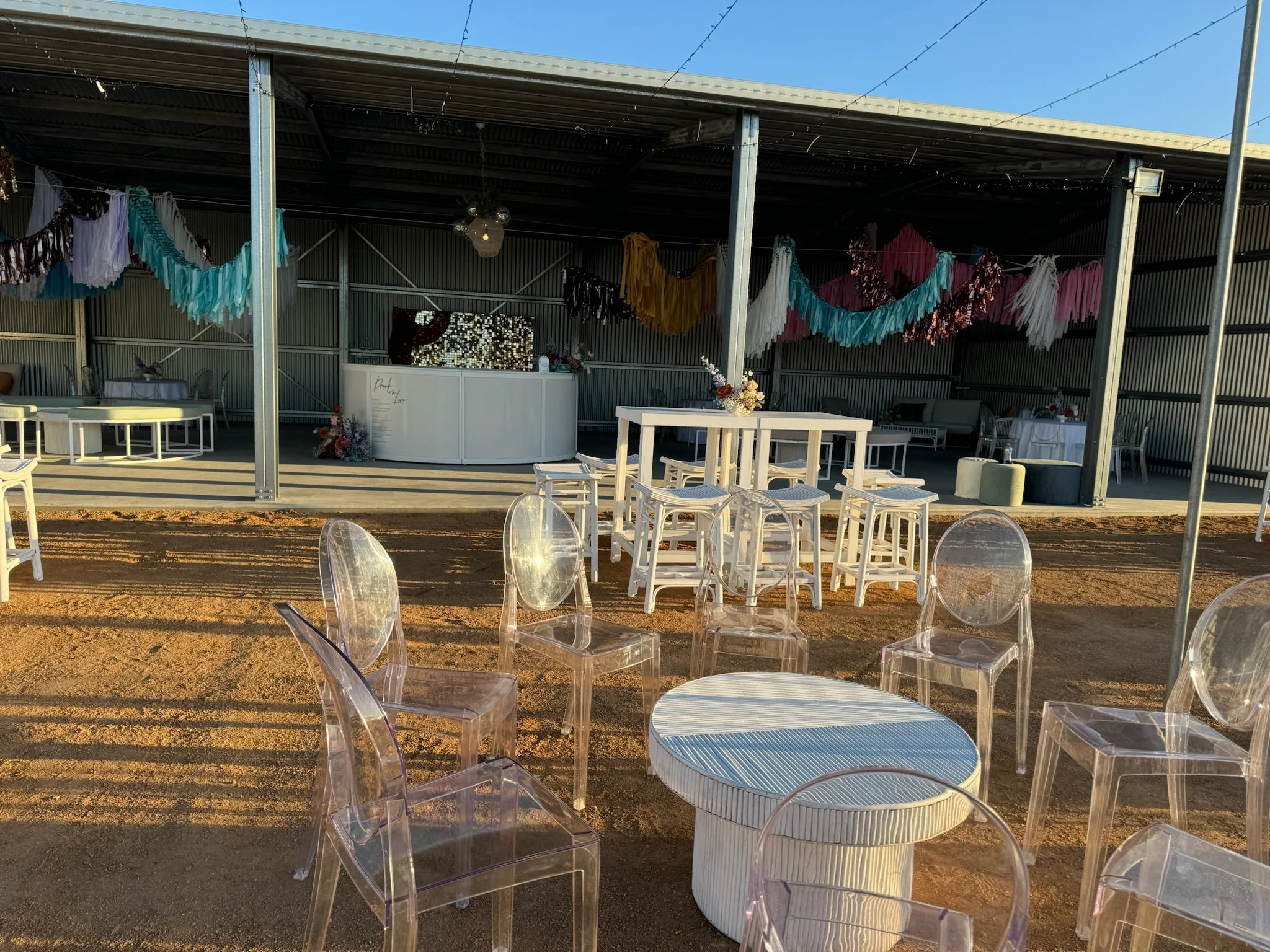 Outdoor event setup with clear plastic chairs and white tables in front of a decorated pavilion featuring colorful hanging tassel garlands and a white bar counter, set on sunny dirt ground.