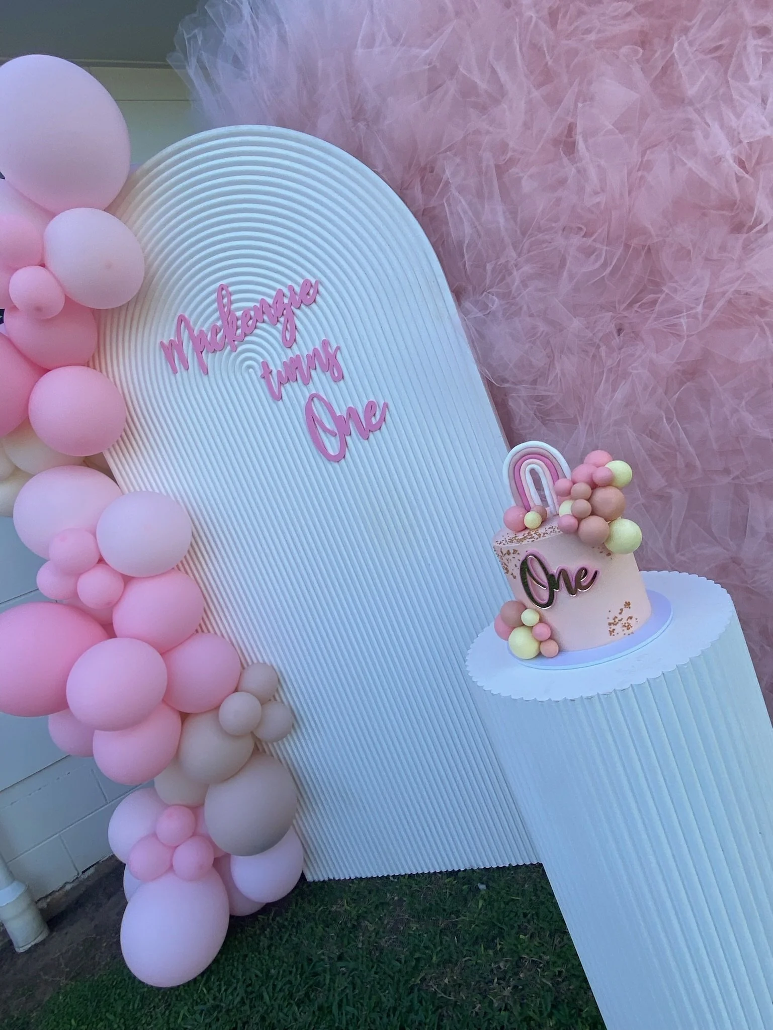 Pink and white balloon arch, a white backdrop with pink text reading 'Mackenzie turns One,' a pink birthday cake with gold accents, a rainbow decoration on top, and pink, yellow, and beige spherical decorations surrounding the cake