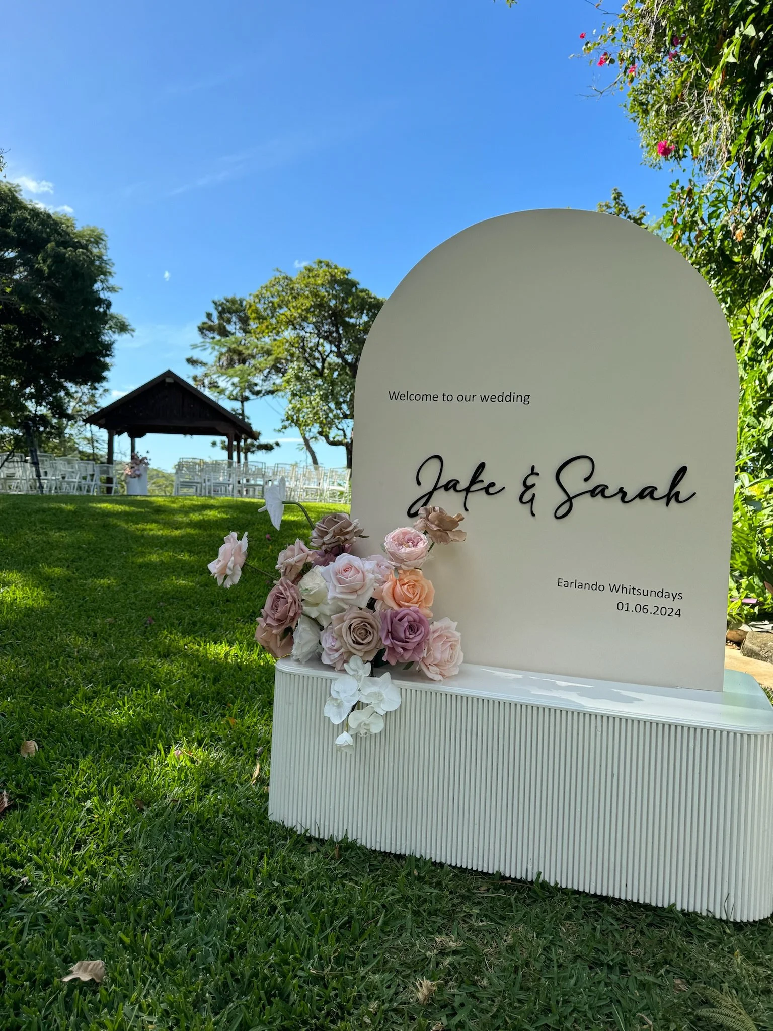 Wedding sign with flowers on a grassy outdoor area, with chairs and a pavilion in the background under a clear blue sky.