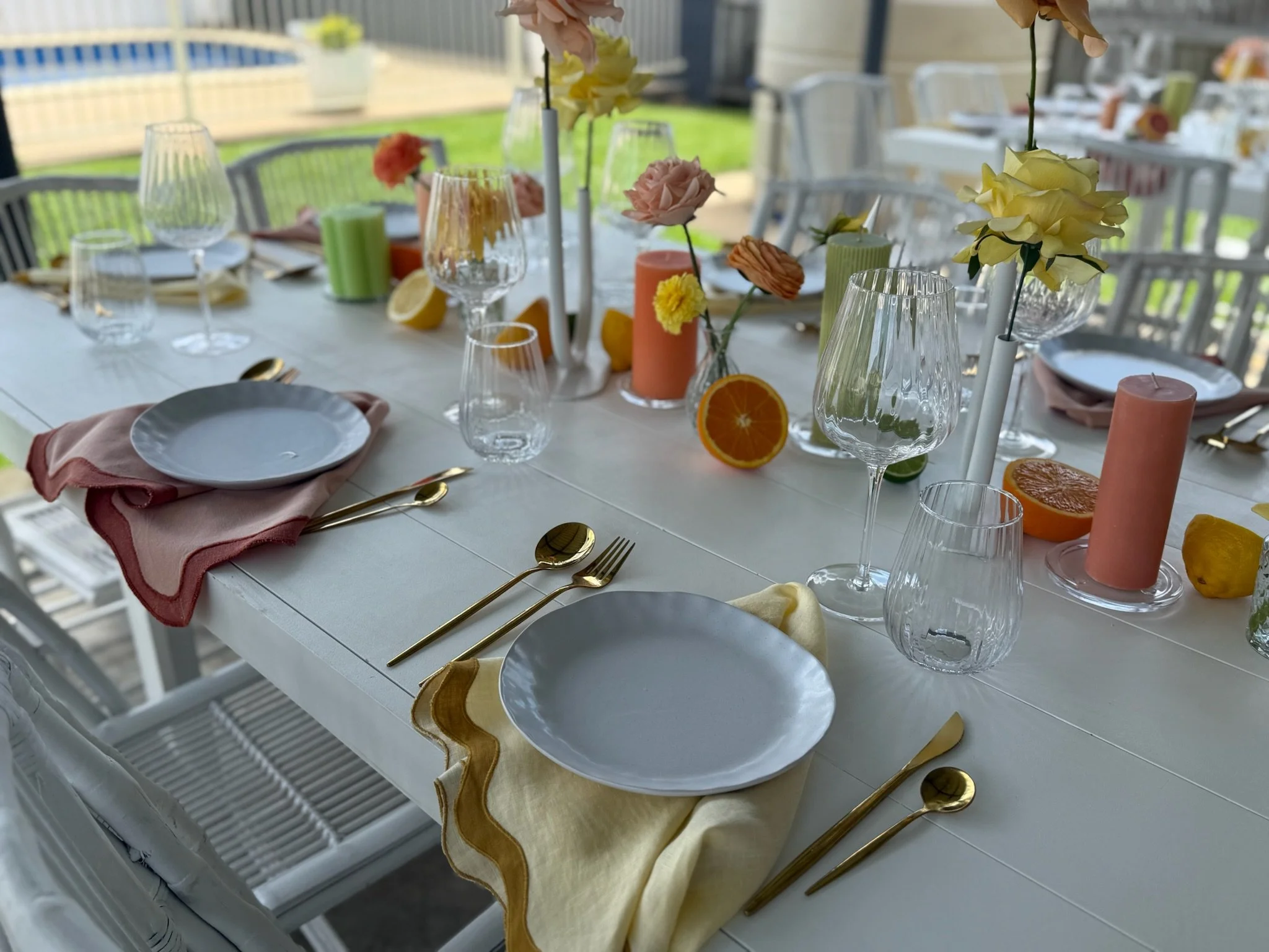 Elegant outdoor dining table decorated with pastel-colored napkins, clear wine glasses, gold flatware, and floral centerpieces featuring pastel flowers and citrus slices.