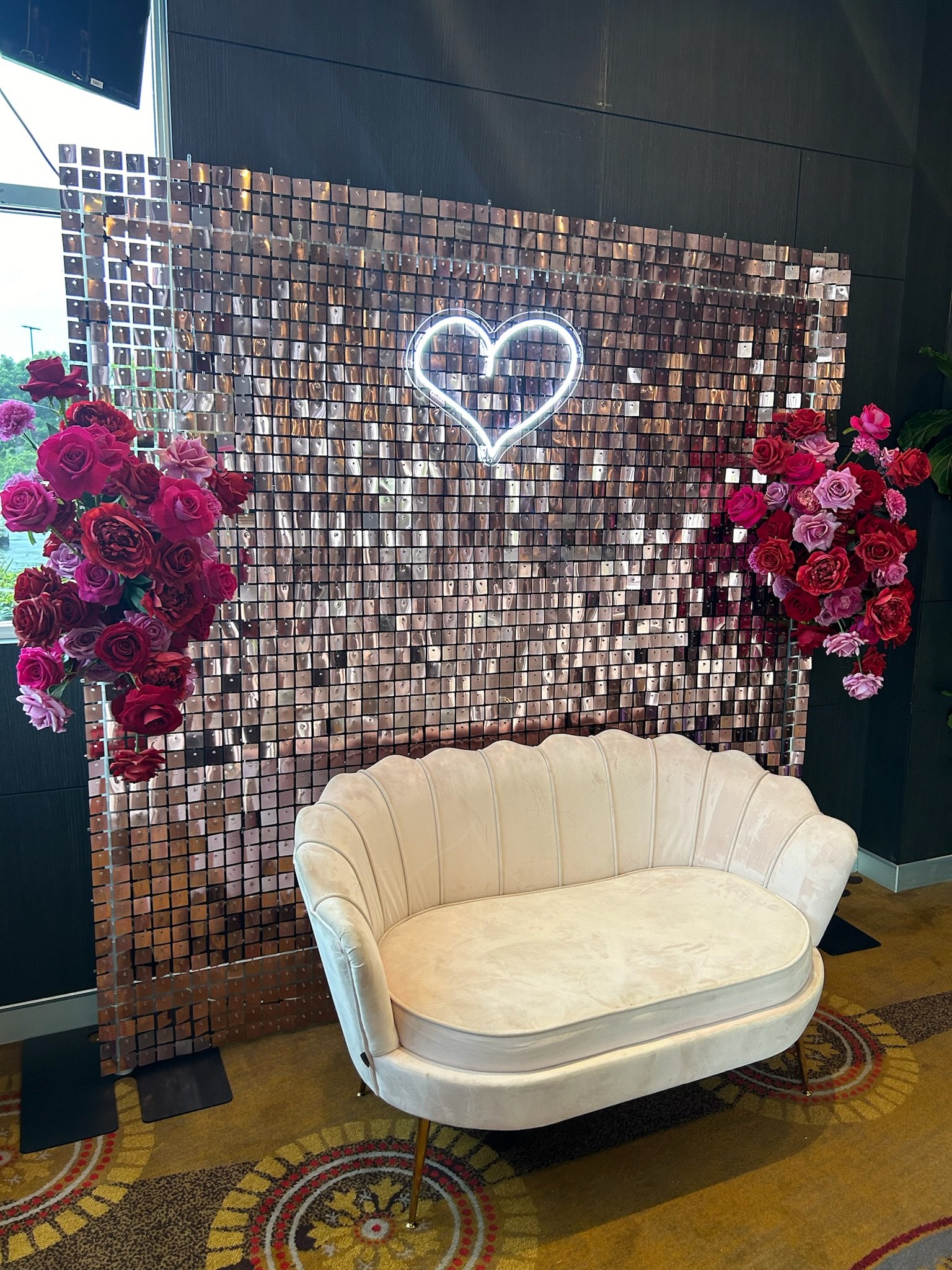 Cream-colored vintage loveseat with a curved backrest and wooden legs, placed in front of a decorative wall with pink and red roses on either side, a heart-shaped neon sign above, and a mosaic tile background.