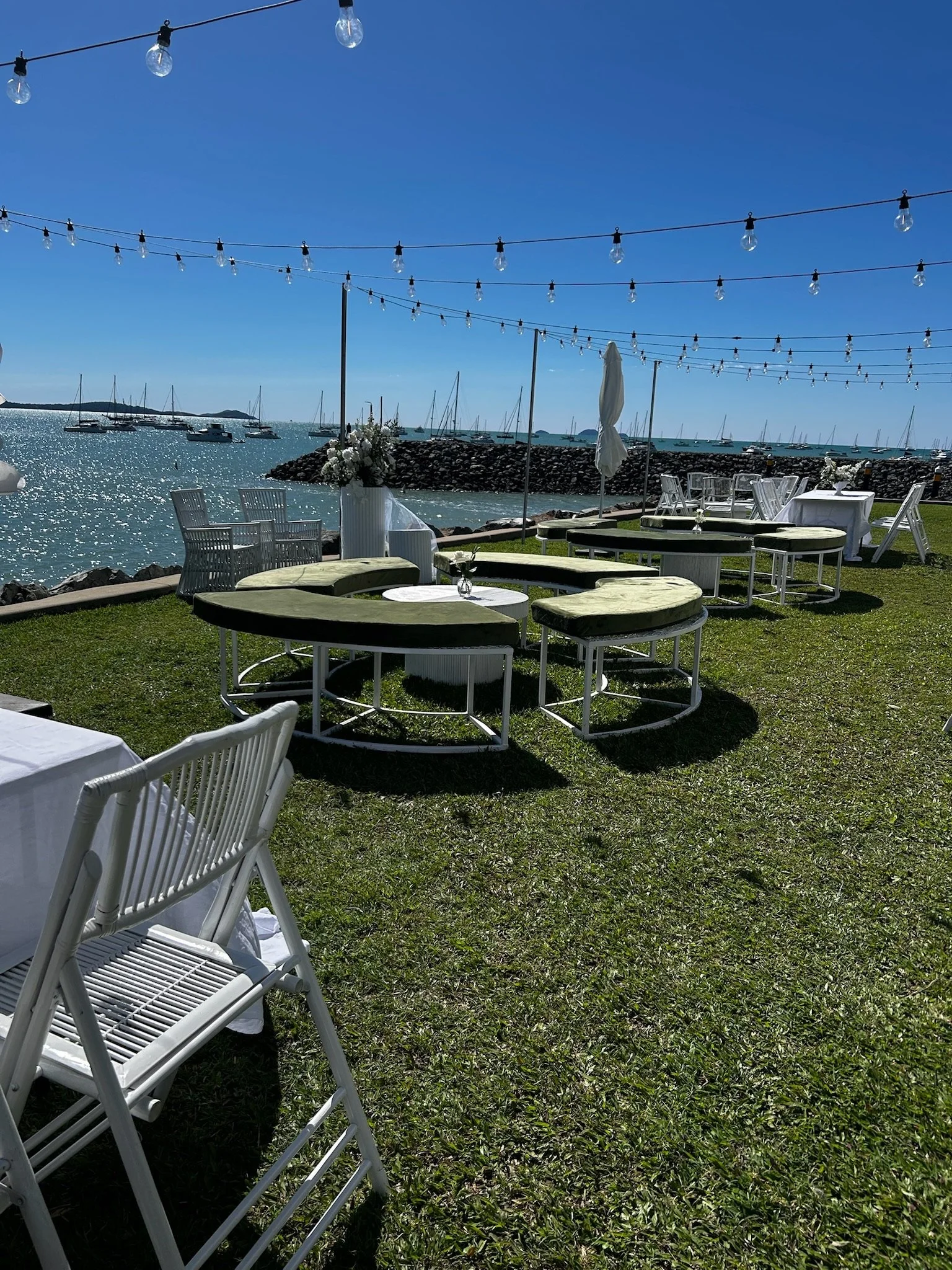 Outdoor event setup by the water with string lights overhead, white chairs, tables, and circular lounging seats, overlooking boats anchored in the marina on a sunny day.