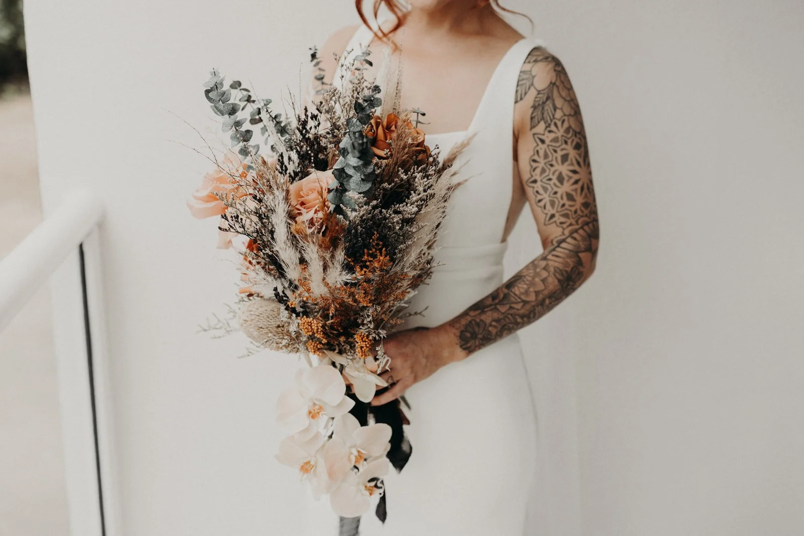 A woman in a white dress holding a bouquet of dried and fresh flowers, with tattoos on her arms and shoulder.