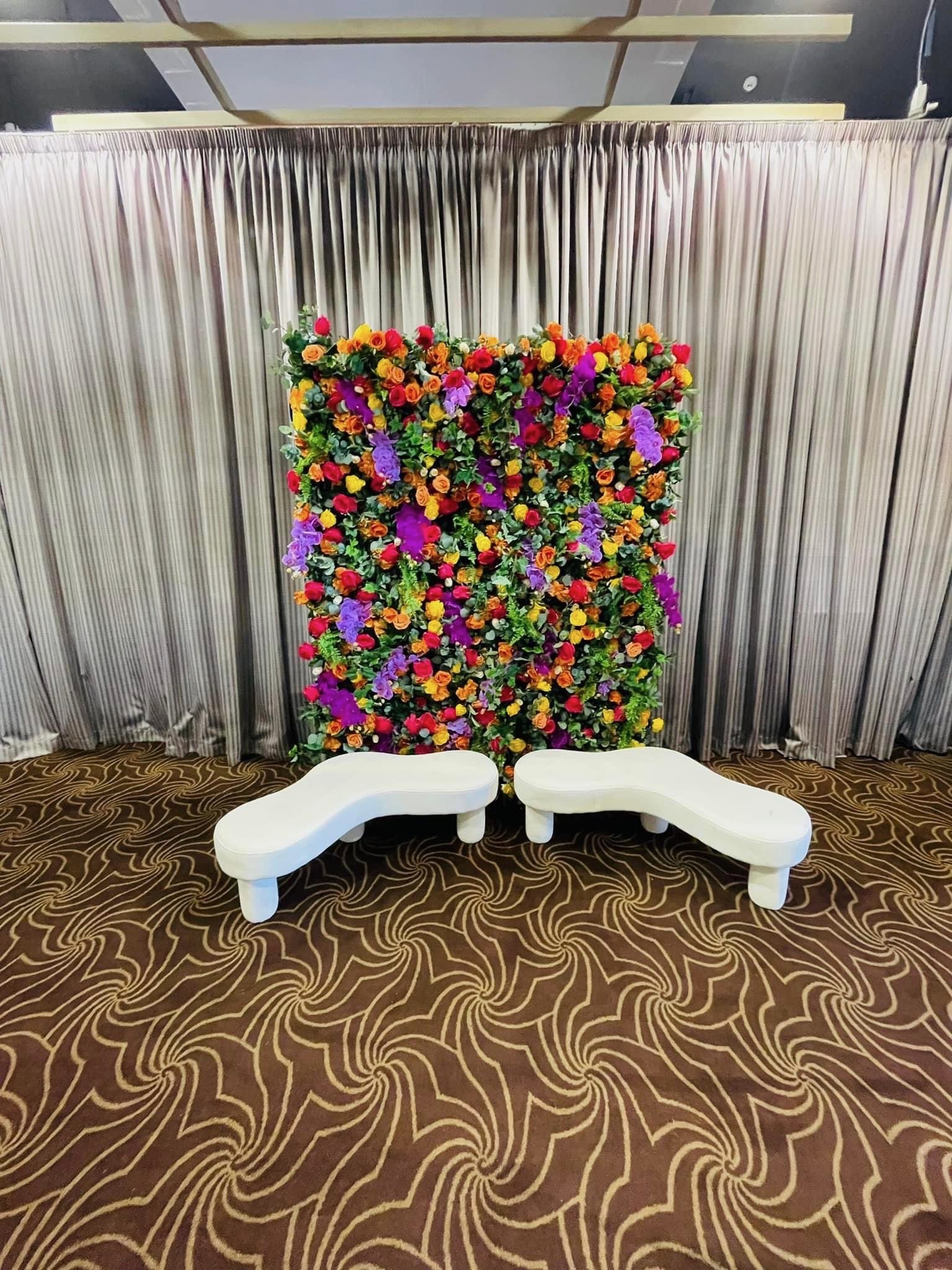 A floral backdrop with colorful flowers, purple flowers, and green leaves, set against silver curtains. In front of the backdrop are two white, curvy benches on a brown patterned carpet.