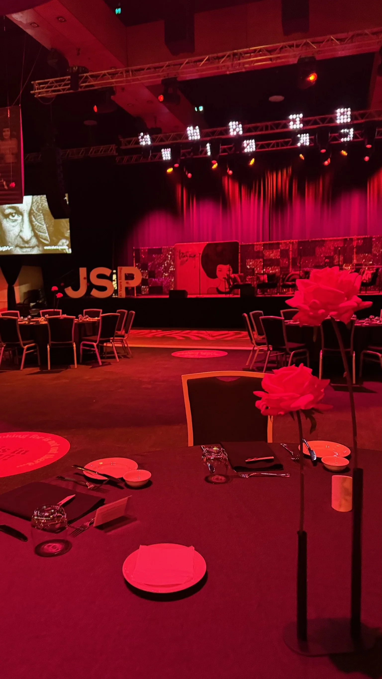 A dimly lit event venue with round tables set for dining, decorated with flowers and glassware, and a stage in the background illuminated with red lighting and 