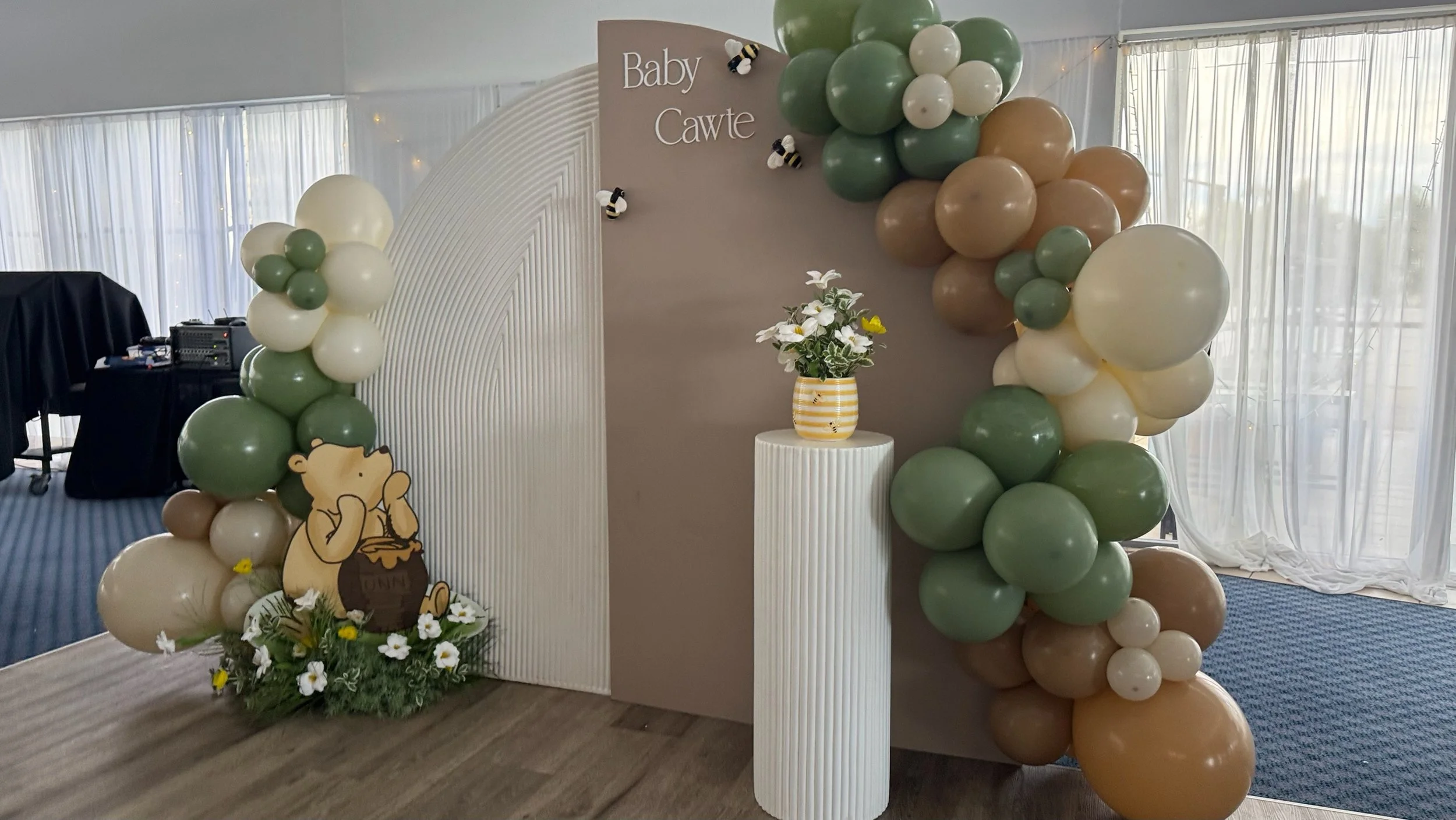 Decorative Baby Cawte event setup with a balloon arch in shades of cream, green, and brown, featuring a Winnie the Pooh figure, flowers, and a flower arrangement on a white pedestal.
