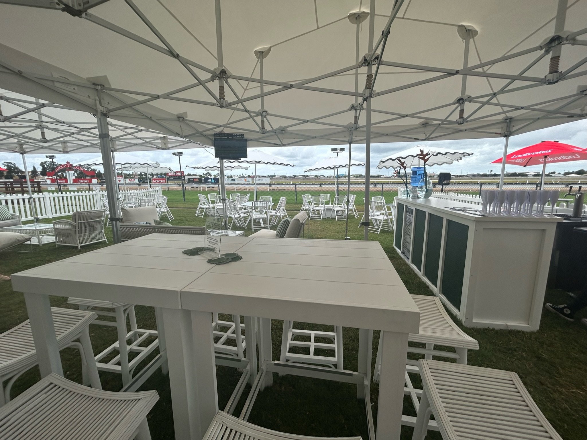 Outdoor seating area at a racecourse with white tables, chairs, and umbrellas, overlooking a racetrack under cloudy sky.