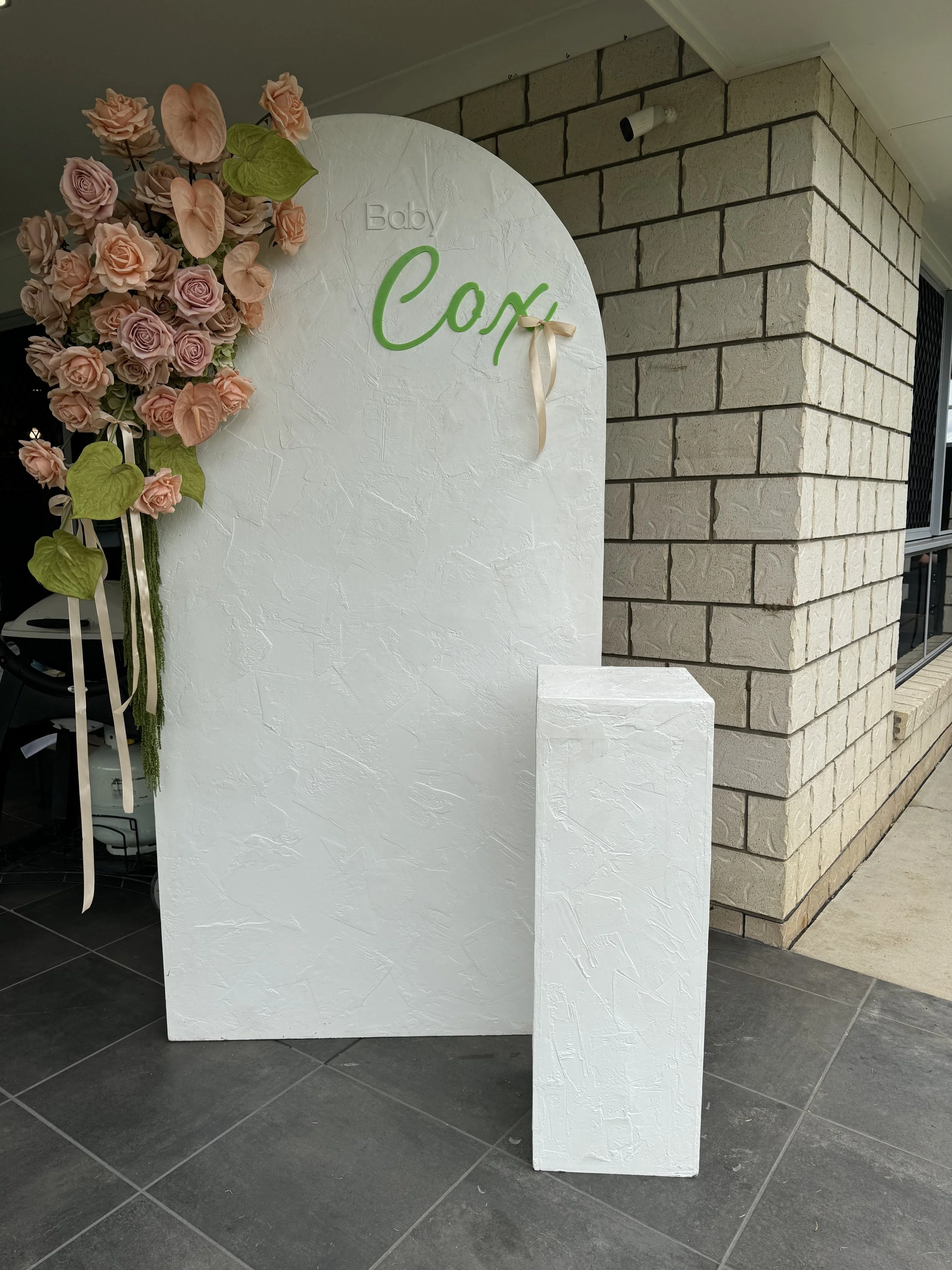 Decorative display for a baby celebration with a white textured backdrop, pink flowers, green leaves, and a green sign that says 'Baby' and 'Con' with a cream-colored ribbon bow.