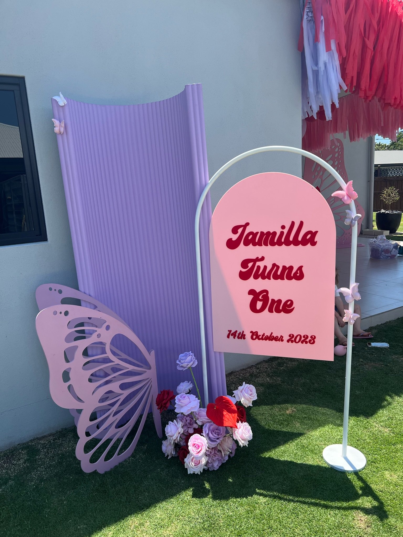 Decorative garden sign for a first birthday celebration with pink and purple butterflies, flowers, and a pink sign reading 'Jamilla Turns One' with the date '14th October 2023'.