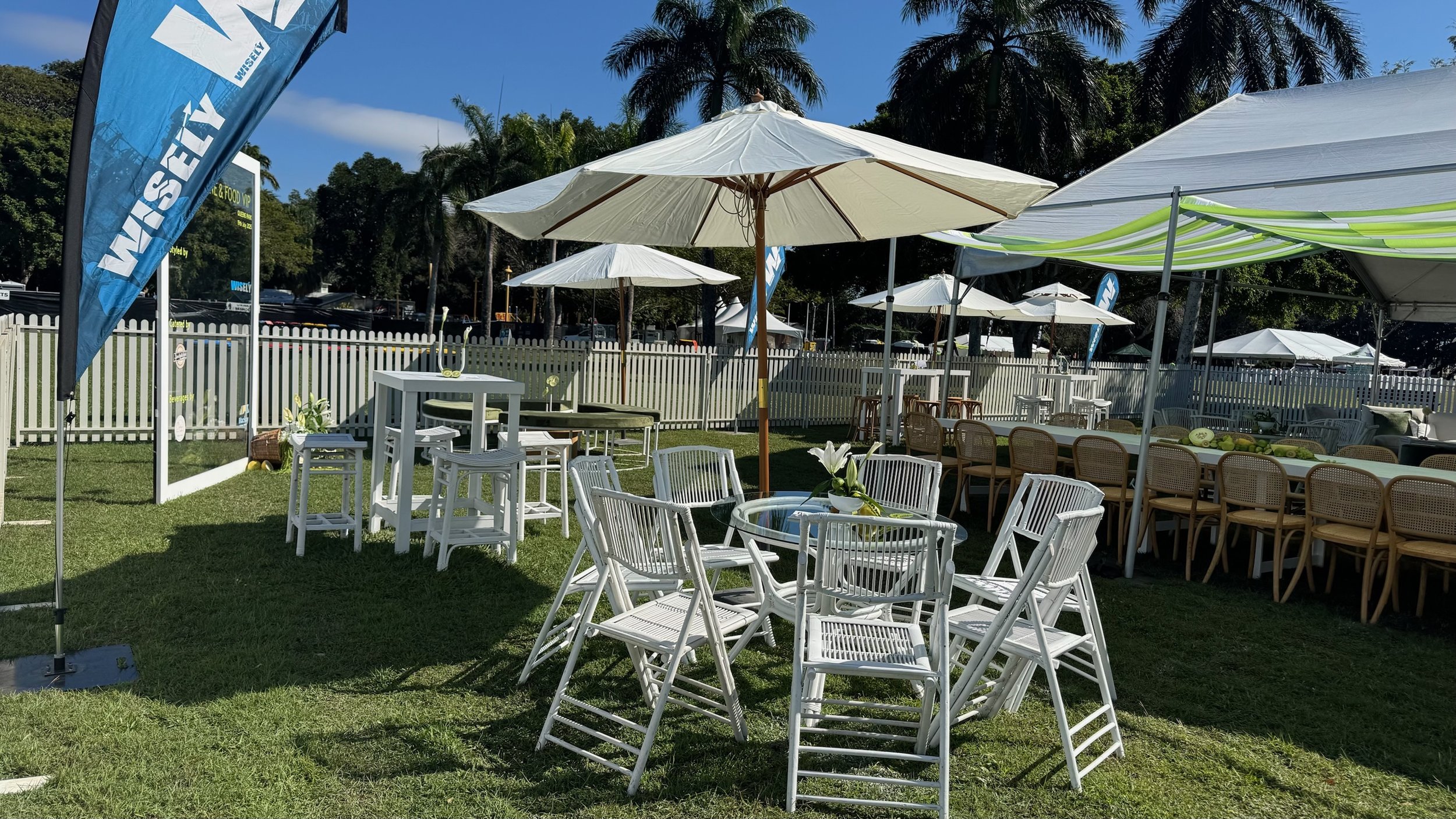 Outdoor event setup with white chairs, tables, umbrellas, and tents on a grassy area surrounded by a white picket fence and palm trees.