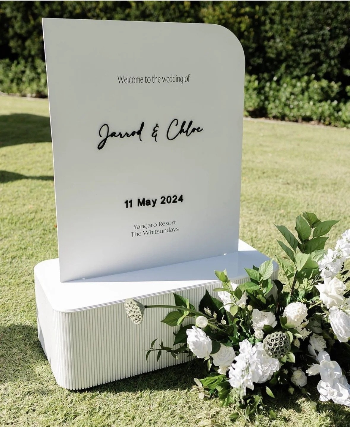 Wedding welcome sign with black text on a white board, reading 'Welcome to the wedding of Jarrod & Chloe, 11 May 2024, Yangaro Resort, The Whitsundays,' on a white stand, decorated with white flowers and green foliage outdoors on a grassy area.