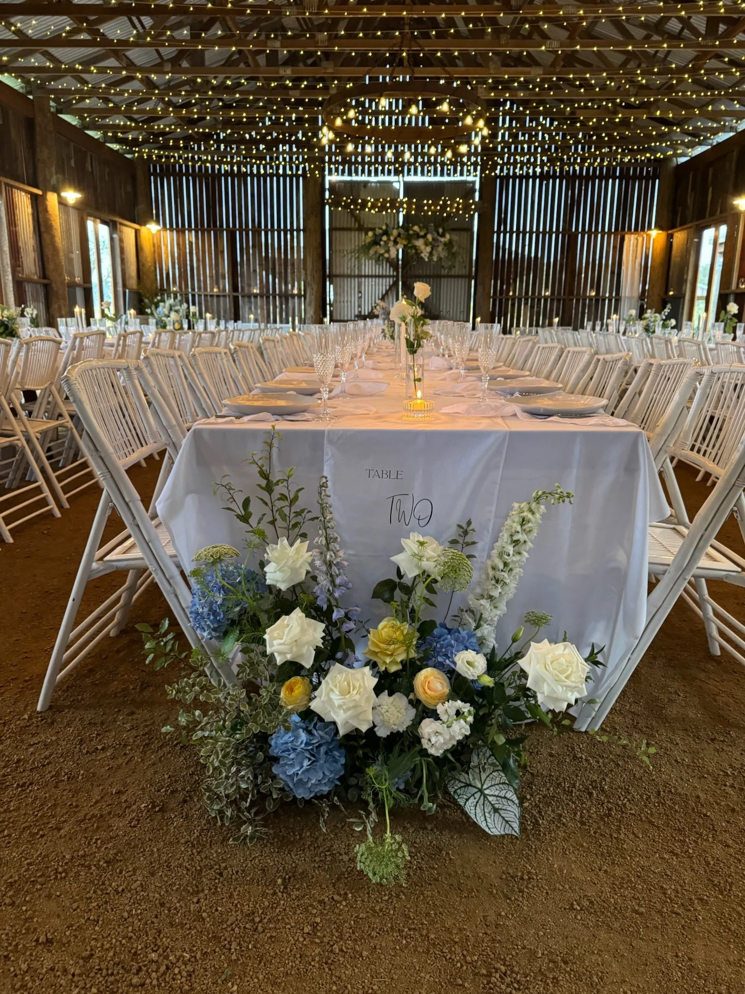 Indoor wedding reception with long white tables, white chairs, floral centerpieces, and string lights hanging from the ceiling, in a rustic barn.