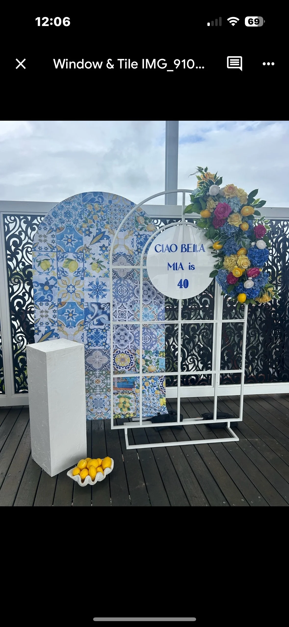 Decorative display for Mia's 40th birthday featuring a white metal frame, colorful flowers, a lemon-themed backdrop, a white pedestal, and a bowl of lemons, with a sign reading 'Ciao Bela Mia is 40'.
