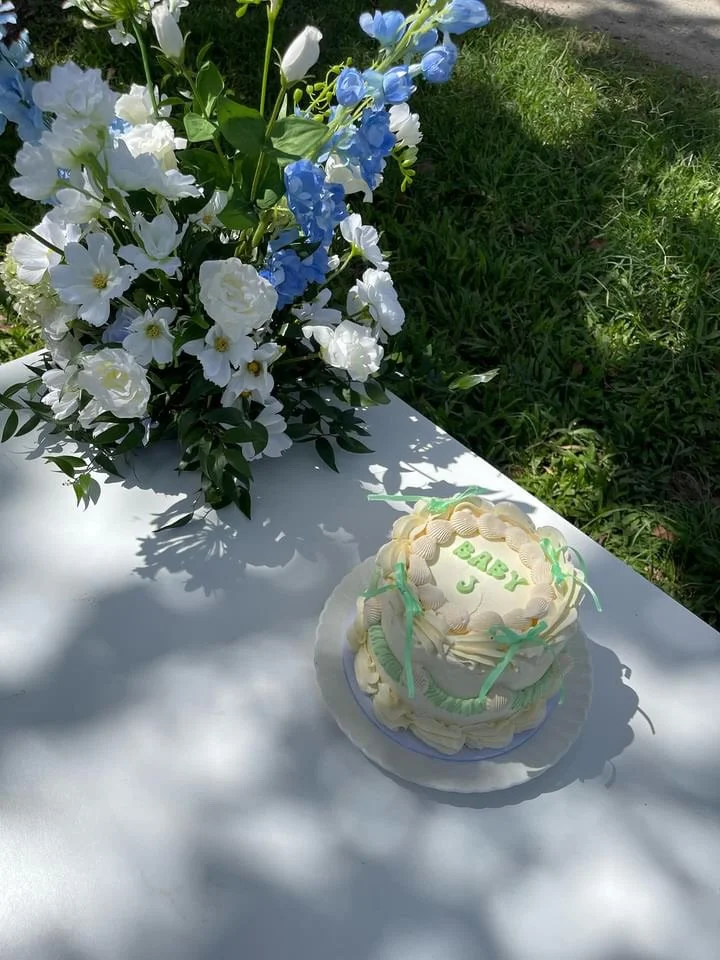 A small, white, two-tiered cake decorated with green ribbons and the words 'Baby J' on top, sitting on a white surface next to a large bouquet of white and blue flowers outdoors on a sunny day.