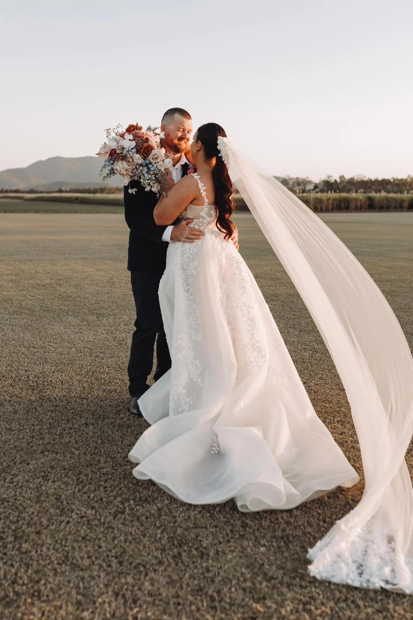 Bride and groom embrace on a grassy field at sunset, with the bride wearing a white wedding gown and veil, and the groom in a dark suit holding a bouquet of flowers.