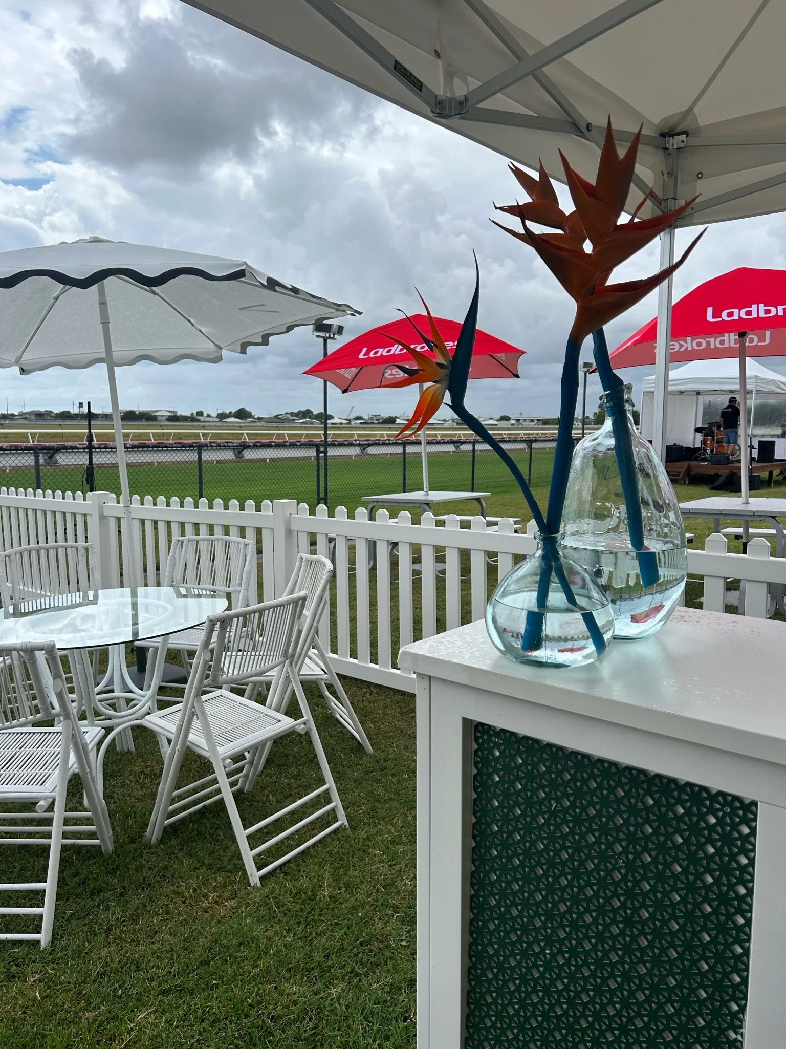 Vase with bird of paradise flowers on white table at outdoor event, white chairs, umbrellas, and race track in background on cloudy day.