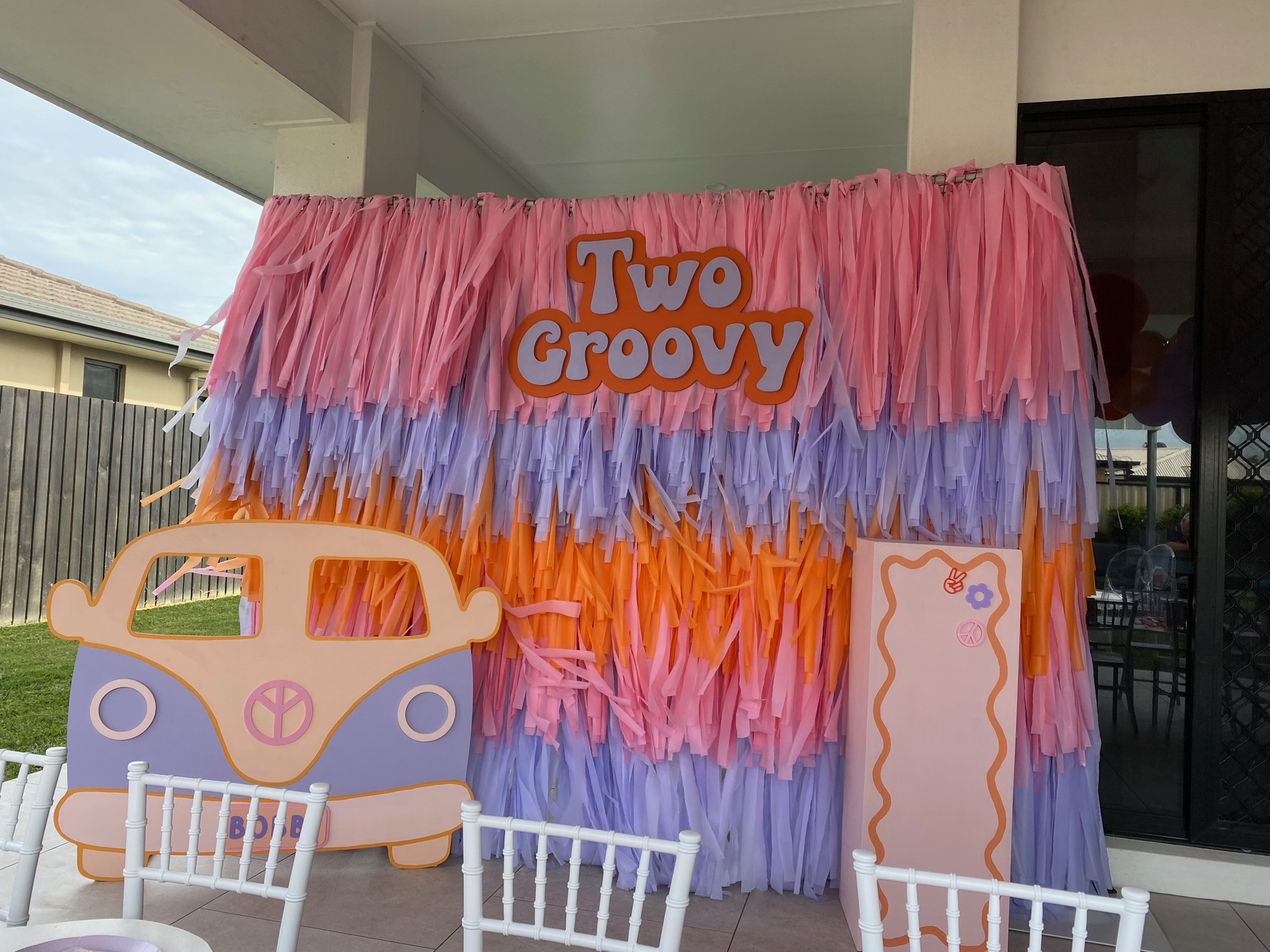 Decorative backdrop with pink, lavender, and orange fringe, featuring the phrase 'Two Groovy' in orange letters, with a pink and blue cartoon camper van cutout, and a pink board with peace and flower symbols for a 1960s or 1970s themed event.