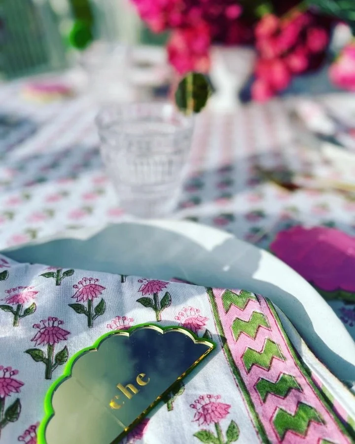 A table setting with a floral cloth, a decorative mirror, a glass of water, and a bouquet of pink flowers in the background.