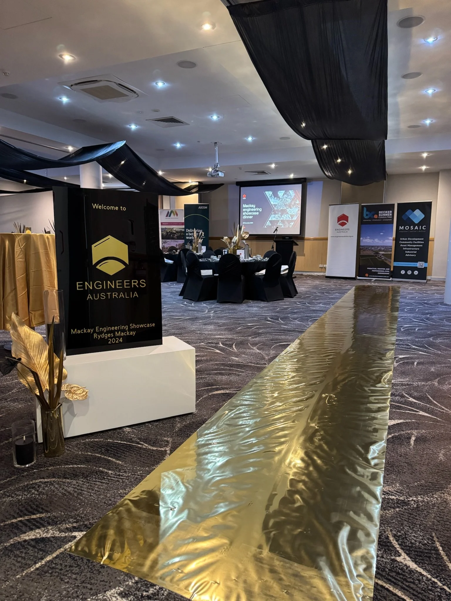 Event space with round tables decorated with black covers and white table settings. There are multiple banners and a stage with a large screen showing presentation slides. A black and gold centerpiece with vases and decorative plants is in the foregr