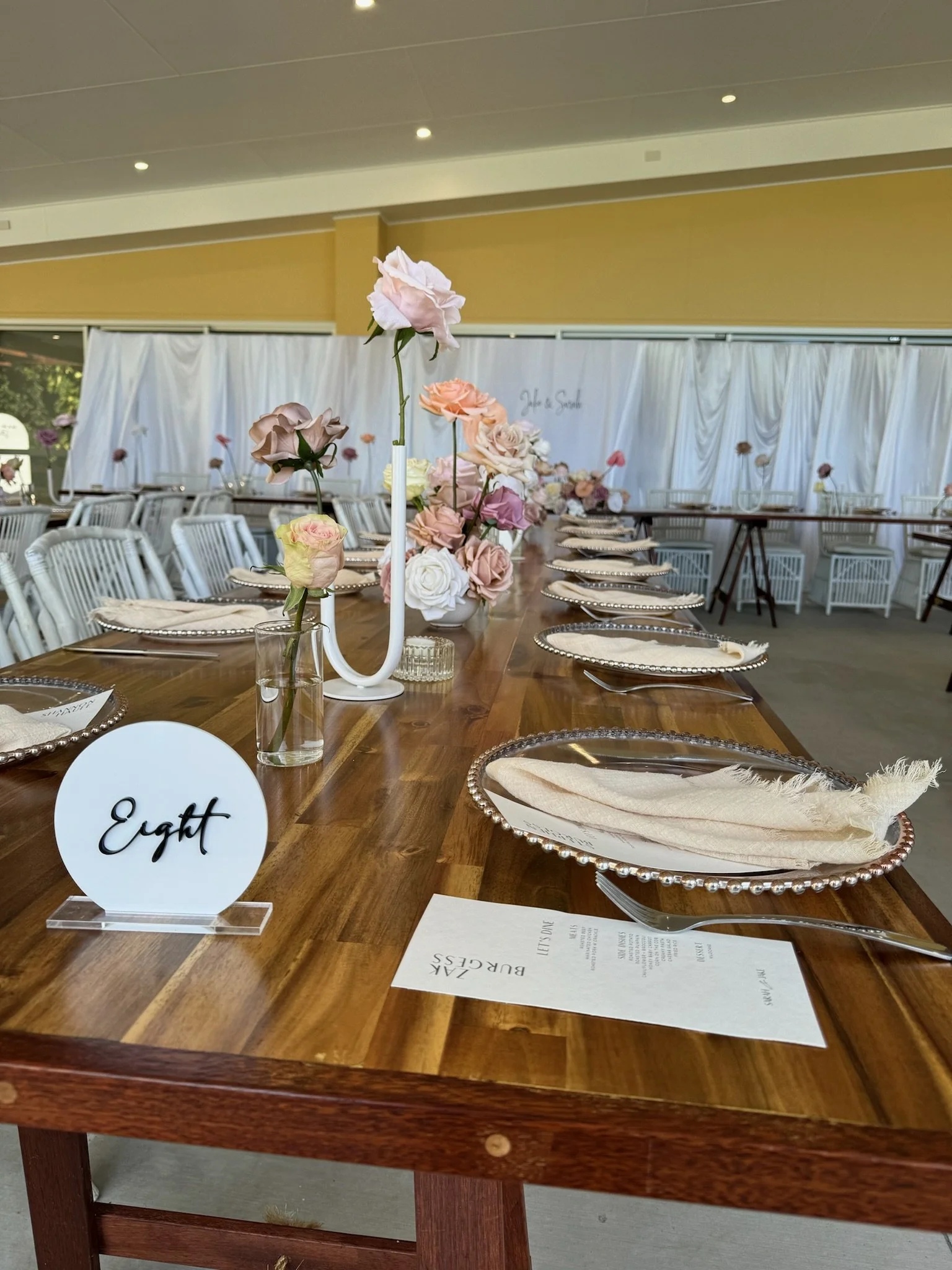Elegant wedding or event table setting with pink and white flowers, silver chargers, and cream napkins, decorated with a white table number sign reading 'Eight'.