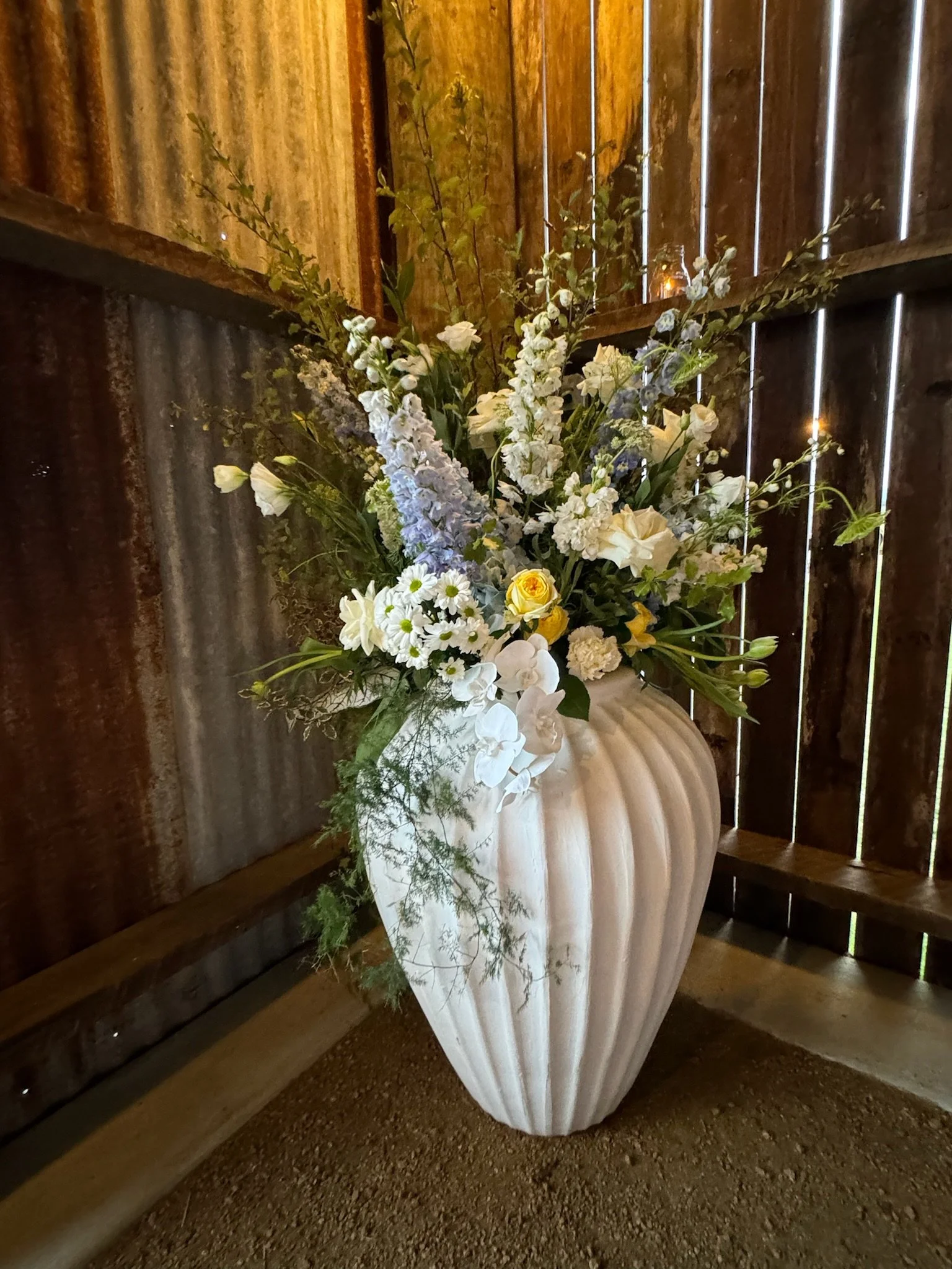 A large beige vase with vertical ridges holding a bouquet of white, yellow, and purple flowers inside a rustic wooden barn with corrugated metal walls.