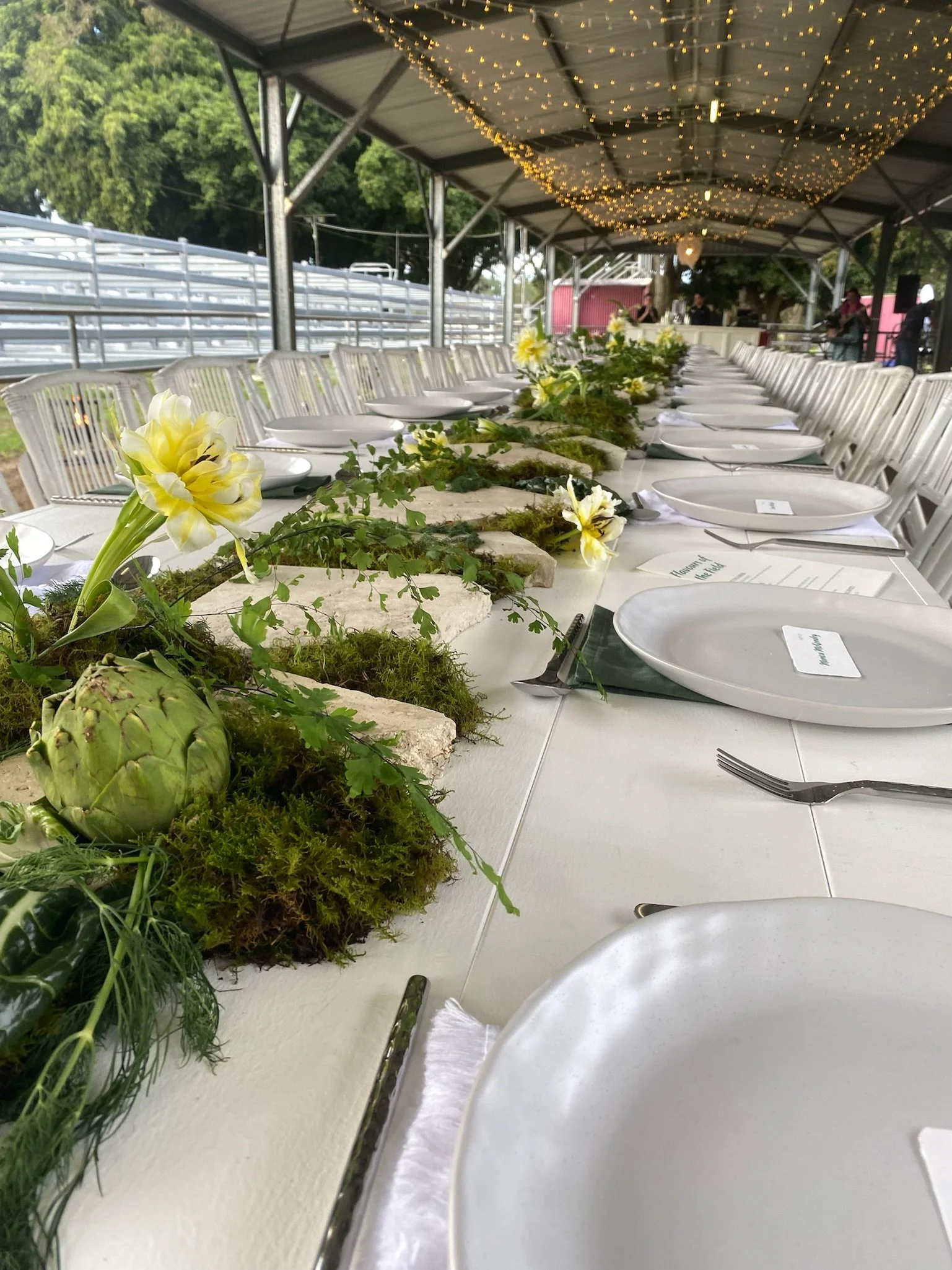 A long decorated tablescape with white tablecloth, white plates, and silverware under a tent with string lights, surrounded by chairs for an outdoor event.