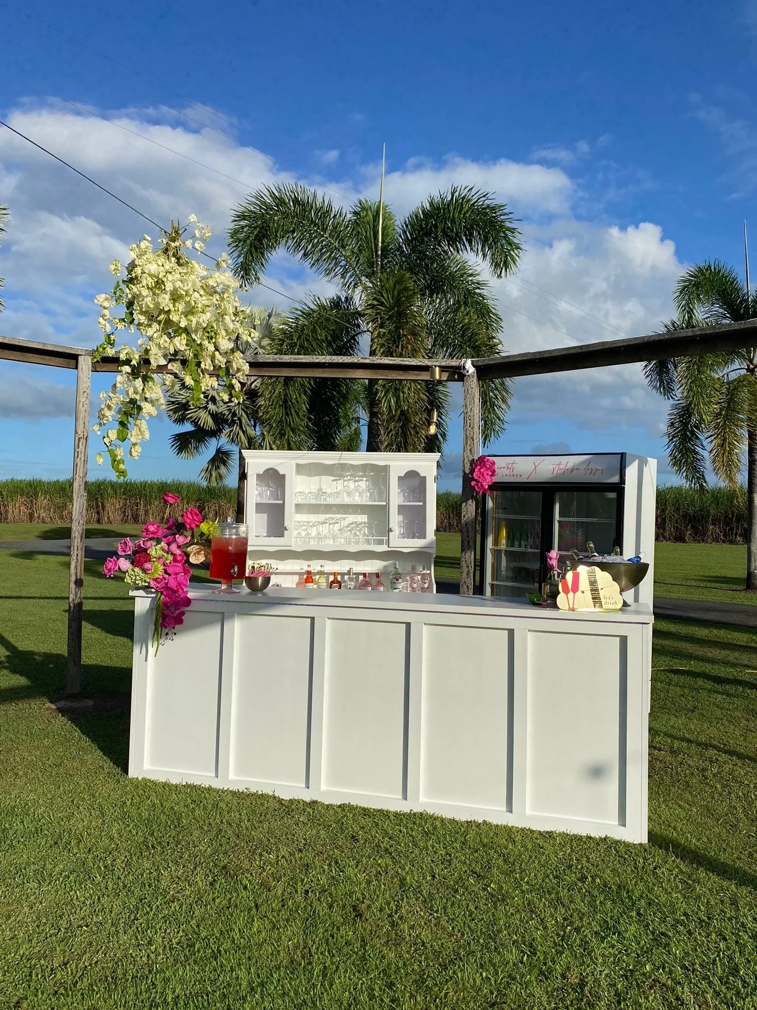 A white outdoor bar setup with a drink dispenser, glassware, and decorative flowers, set against palm trees and a blue sky.