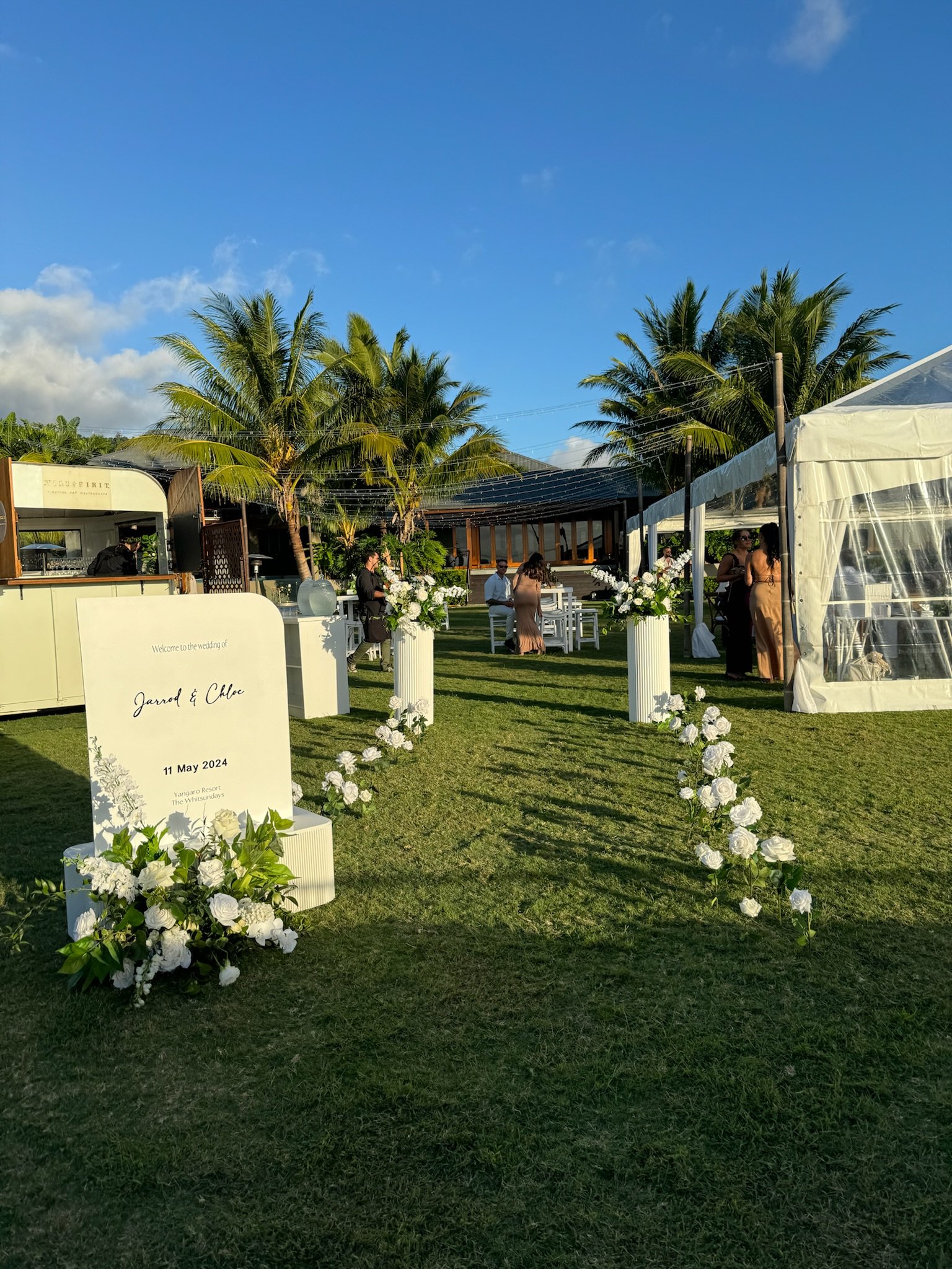 Outdoor wedding setup with white floral decorations, aisle with white roses, large sign welcoming guests for Jared and Chloe's wedding on May 11, 2024, at a tropical resort with palm trees and a blue sky.