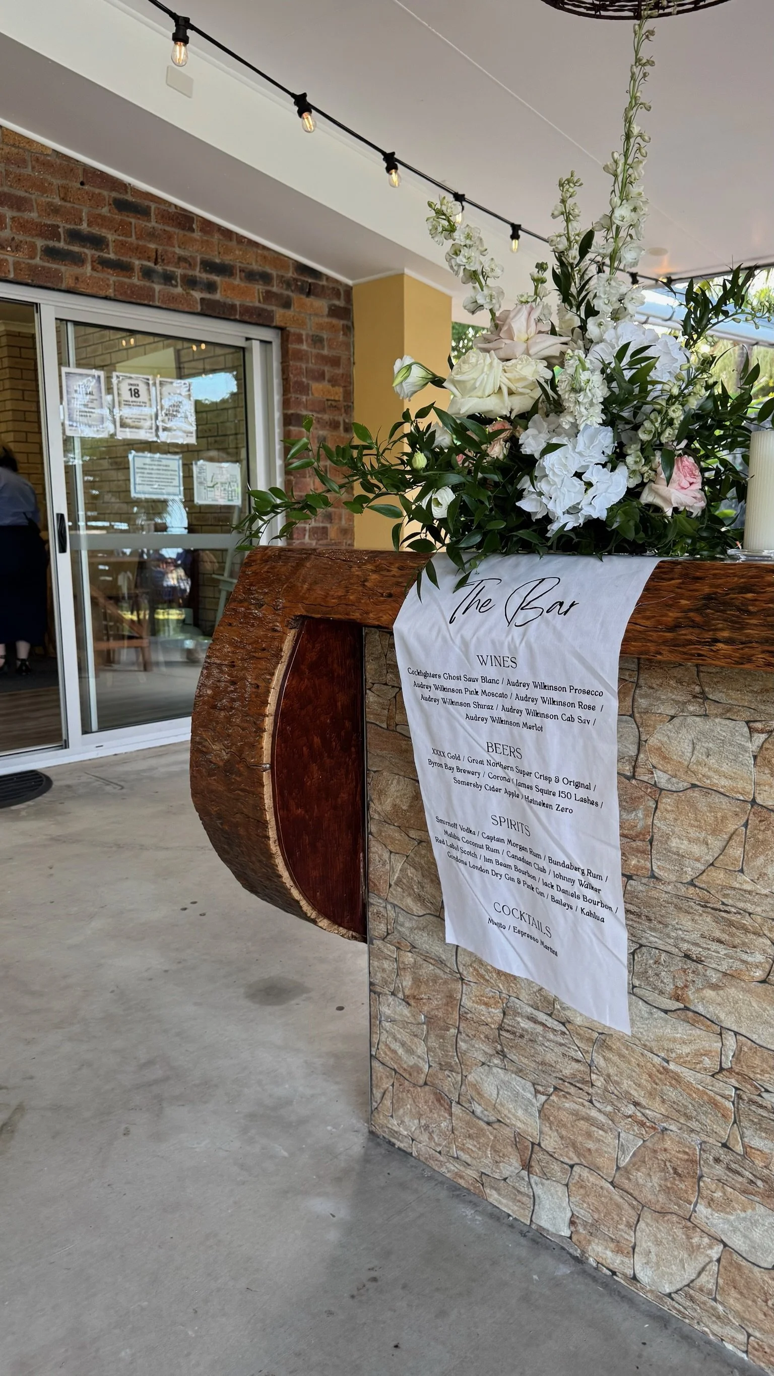 A rustic bar stand with a floral arrangement and a menu listing wines, beers, spirits, and cocktails, located outside a building with a brick wall and a sliding glass door.