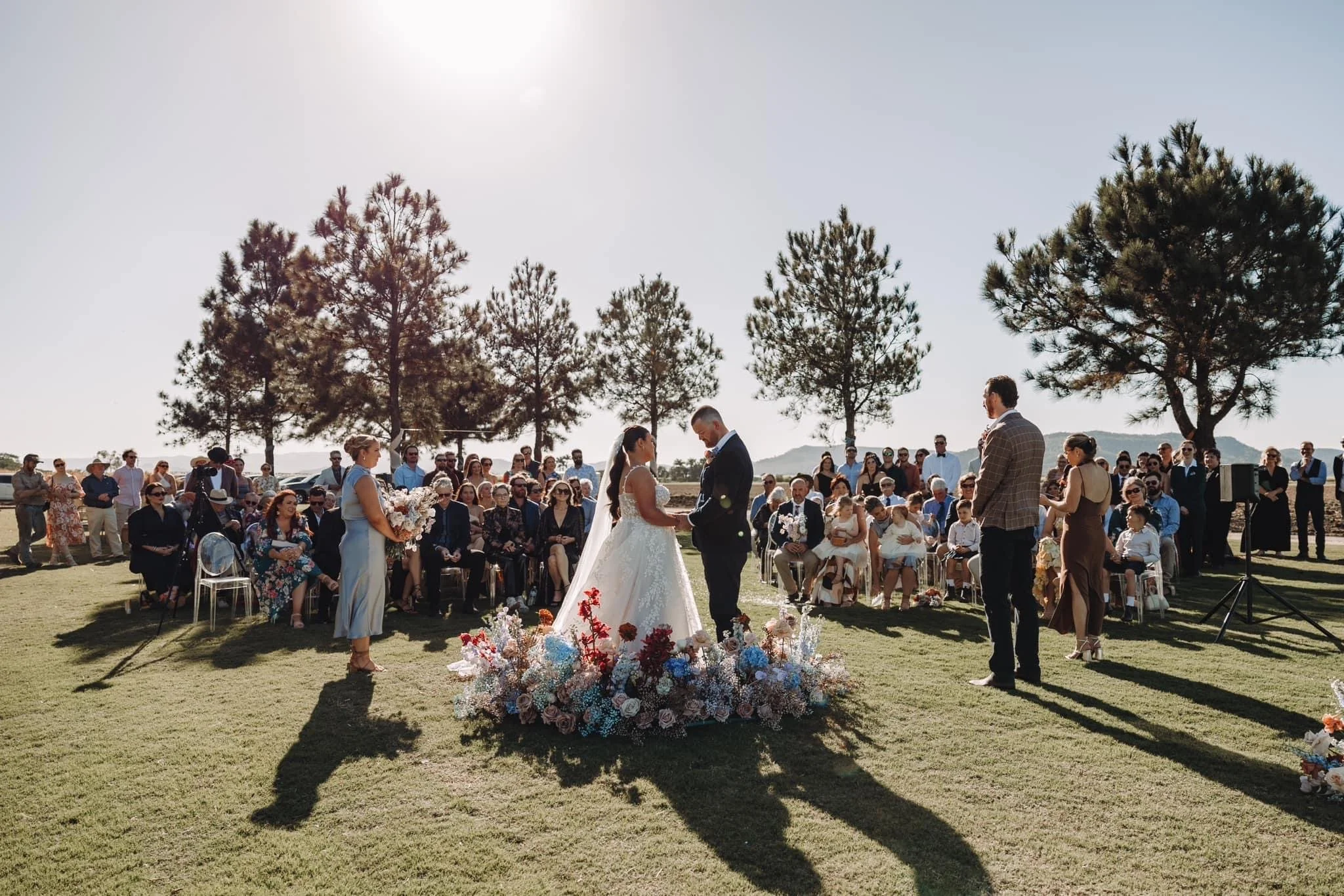 A wedding ceremony outdoors with the bride and groom standing together at the altar, surrounded by floral arrangements. Guests are seated and standing, watching the ceremony in a scenic setting with trees and clear sky.