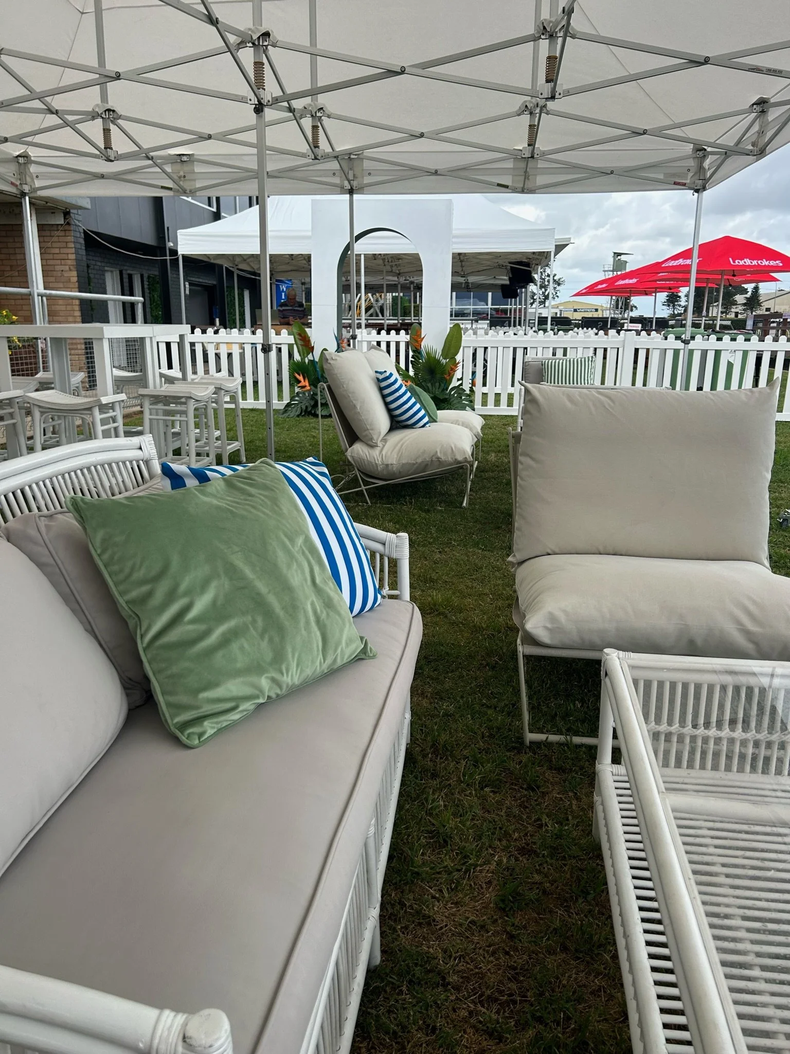 Outdoor seating area with white wicker furniture, cushions, and pillows on a grassy lawn under a large white canopy. In the background, there is a white decorative arch, a white picket fence, and a red umbrella.