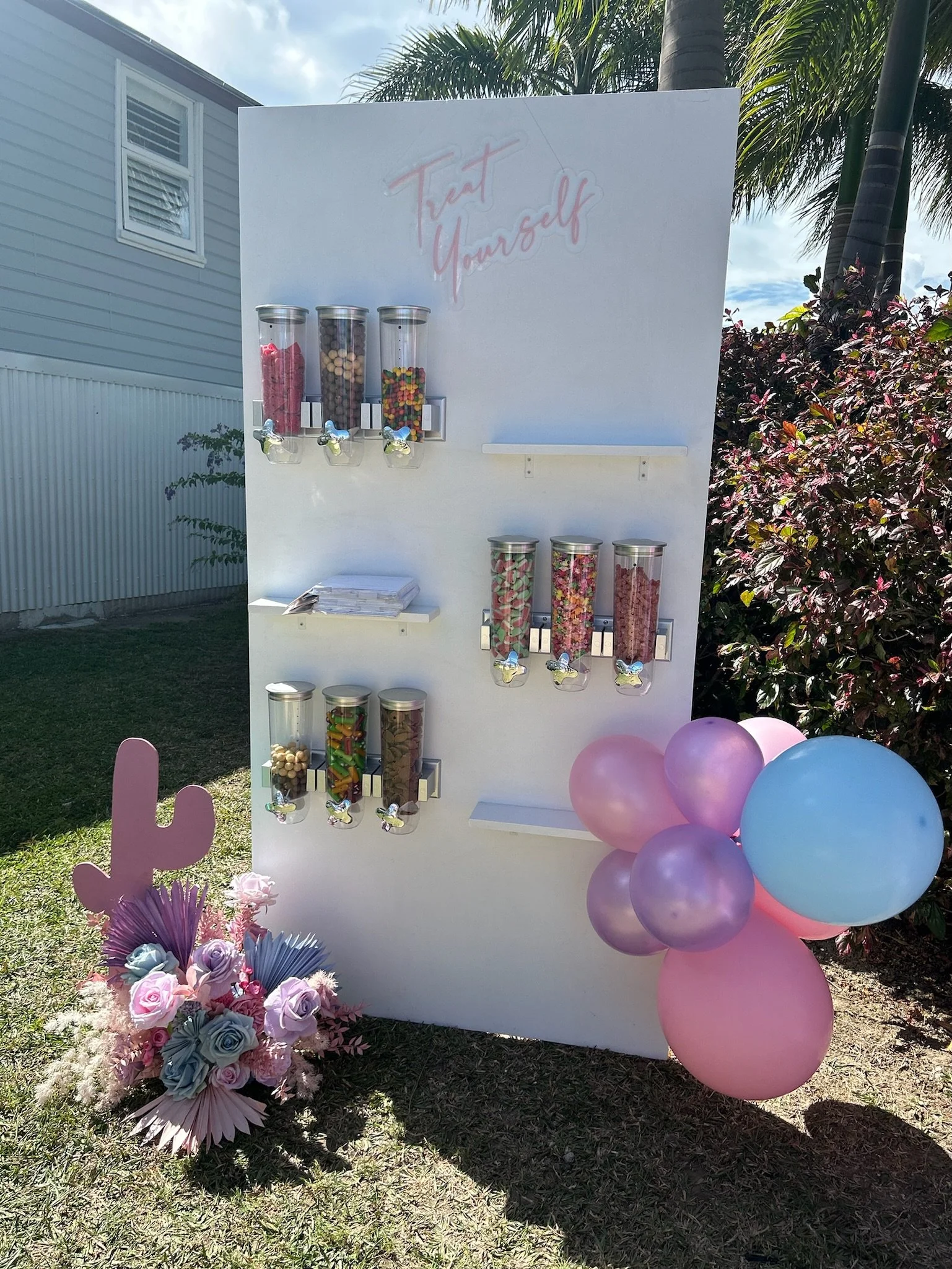 Candy dispenser stand with pink, purple, and blue balloons, a floral arrangement with roses and succulents, and a sign that reads "Treat Yourself" in neon-style lettering, set outdoors on grass with a house and palm trees in the background.