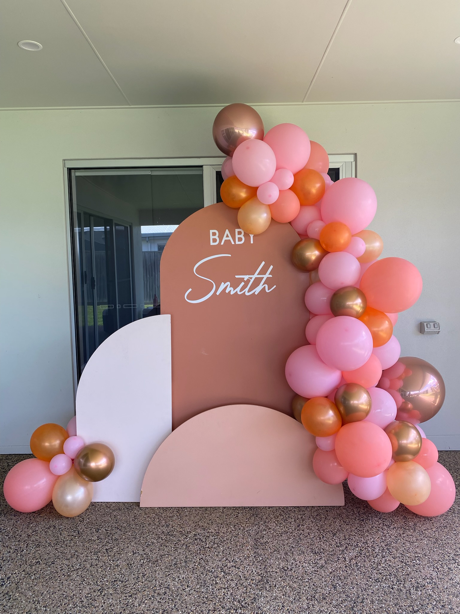 Decorative backdrop for a baby shower or celebration with pink, peach, and gold balloons arranged around a pastel-colored backdrop that reads 'Baby Smith'.