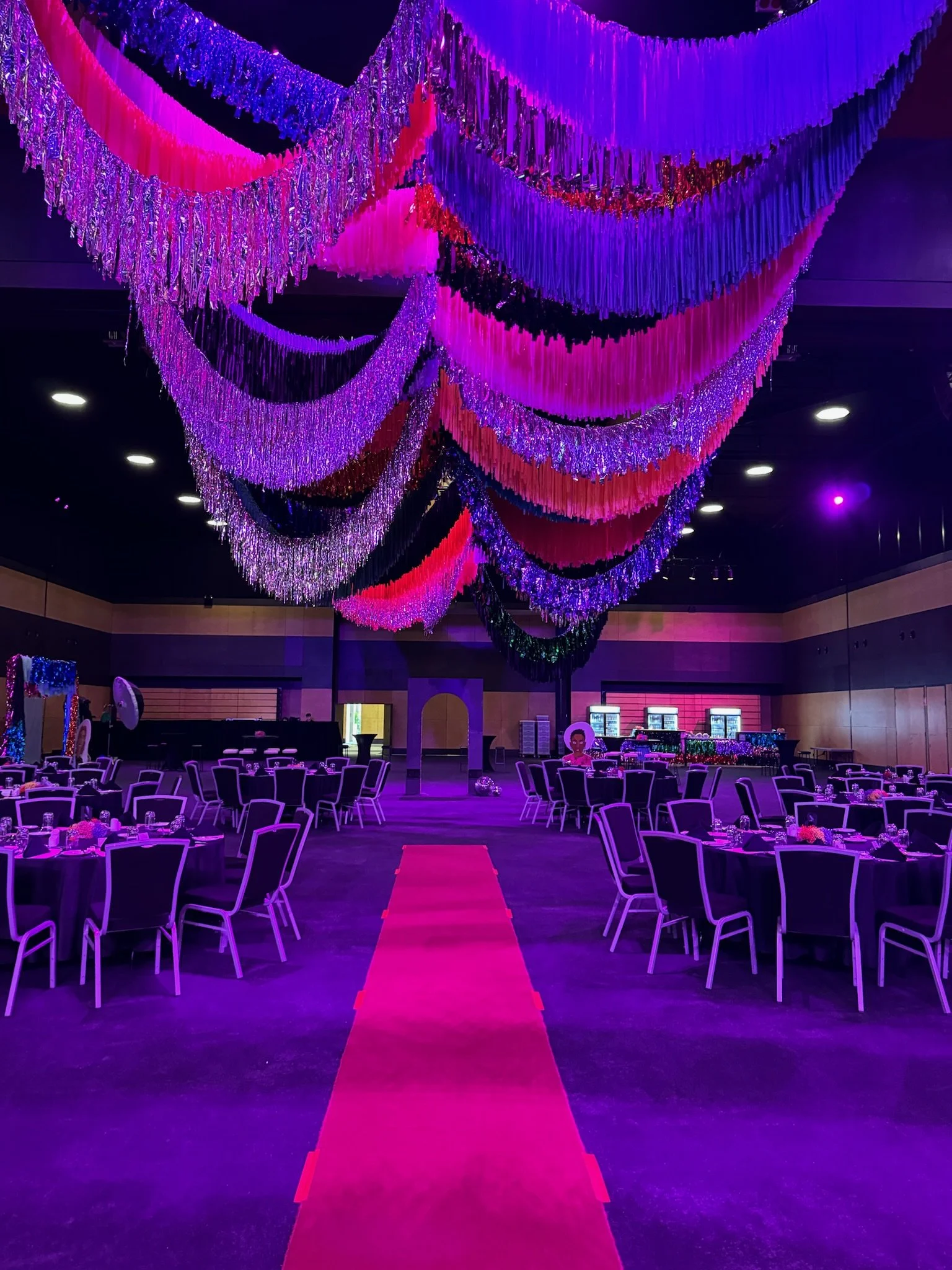 Decorated event hall with purple, pink, and red hanging streamers, round tables with chairs, and a pink carpet aisle in the center.