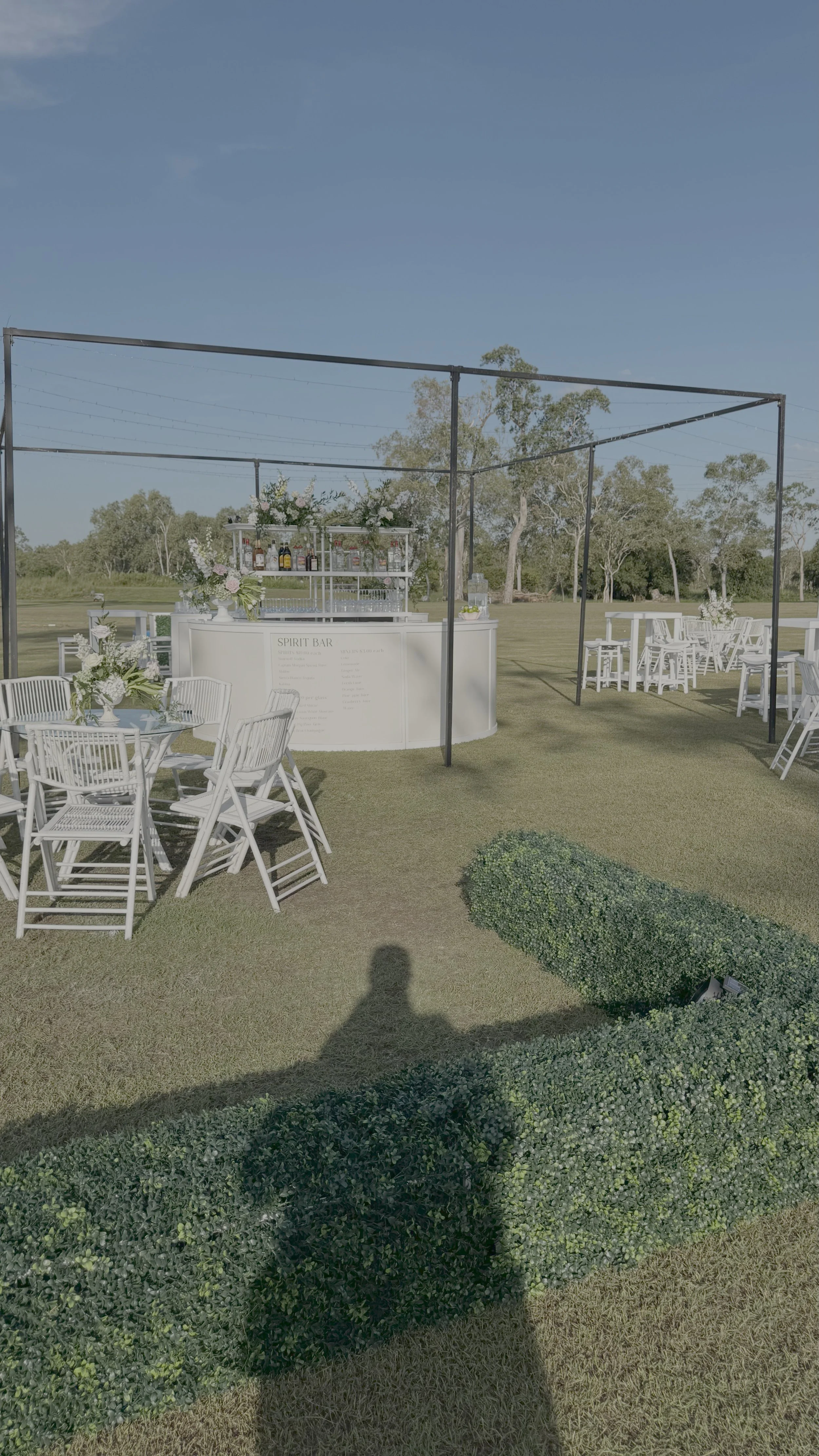 Outdoor wedding or event setup with white chairs and tables, a bar or refreshment stand under a black metal frame, and trees and open skies in the background.