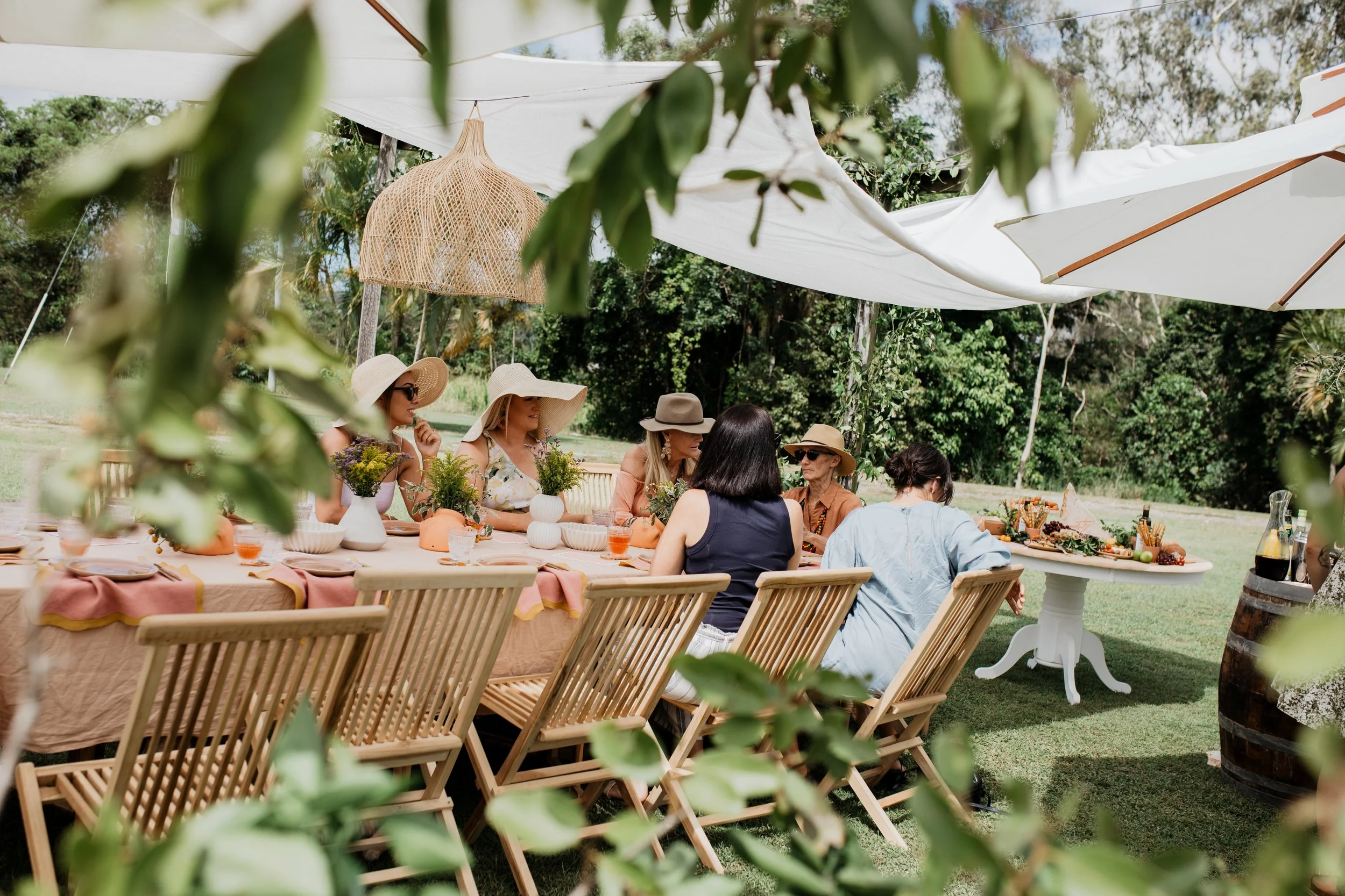A group of women are sitting around a long table outdoors, enjoying a gathering under large white umbrellas, with greenery and trees in the background.