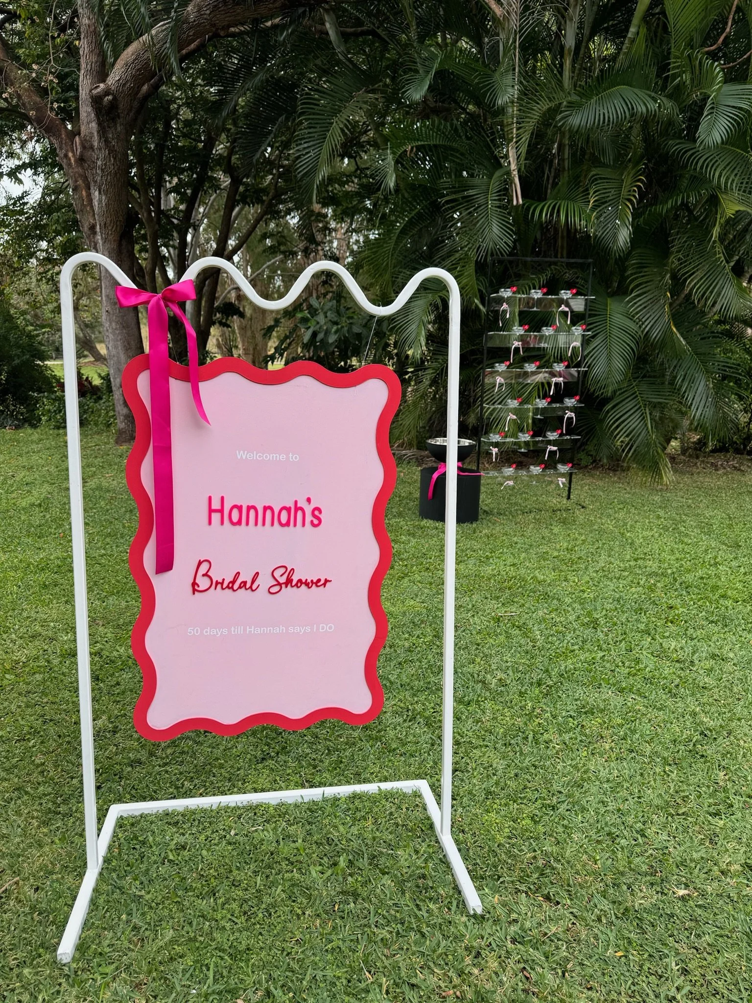 Outdoor bridal shower sign with pink and red decorations, surrounded by green foliage.