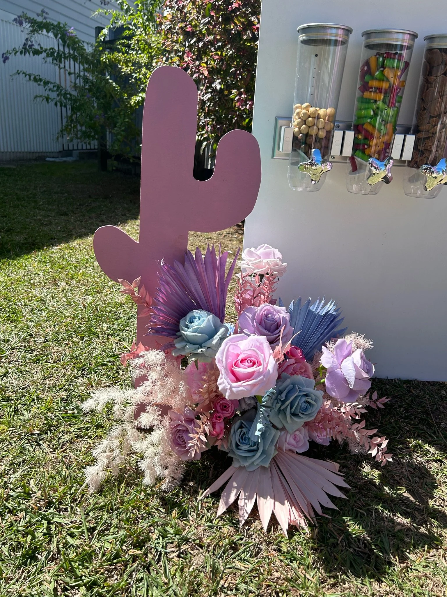 Decorative pink cactus-shaped sign surrounded by pastel-colored flowers, including roses, against a grassy outdoor background with a white cereal station dispensing snacks.