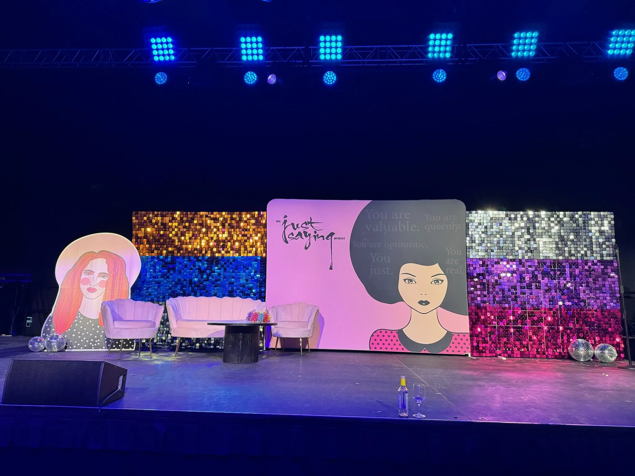 Stage setup with purple and pink couches, a table with colorful decorations, and colorful sequin backdrop panels. There are illustrated portraits of women, stage lights overhead, and disco balls on the floor.