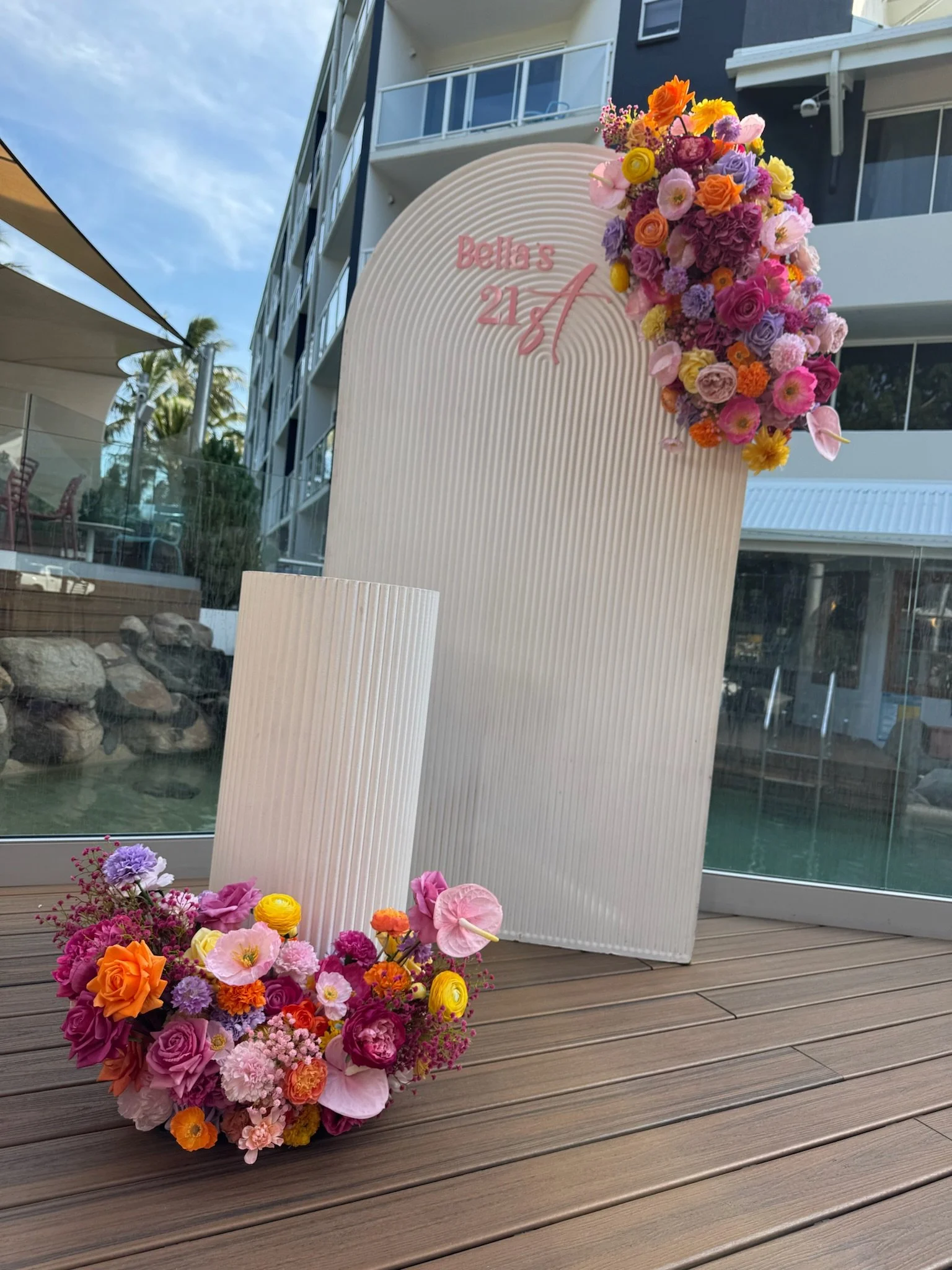 Decorative floral display with pink, purple, orange, and yellow flowers on a wooden deck, with a white backdrop and sign that reads 'Bella's 21st' in pink lettering, at an outdoor venue.