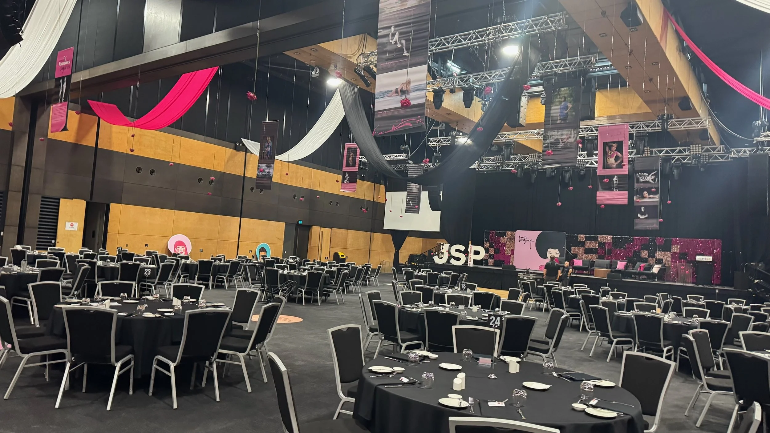 Event hall decorated with black, white, and pink fabric drapes, hanging banners, and stage with large letters 'JSP' and a pink-themed backdrop. Multiple round tables with black tablecloths and chairs are set up for dining, awaiting guests.